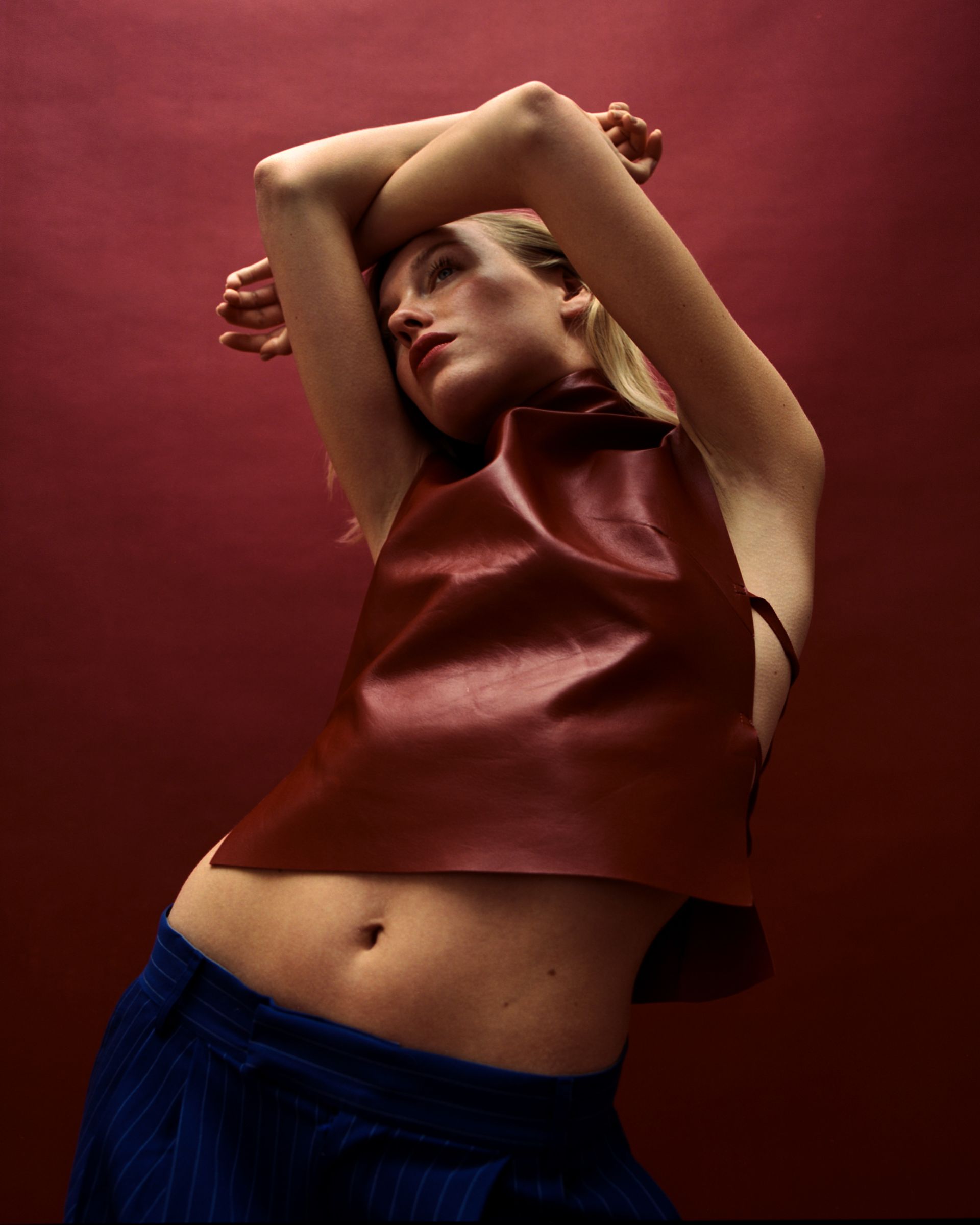 Frau in burgunderfarbenem Lederoberteil und blauen Hosen, die Arme erhoben, vor einem kastanienbraunen Hintergrund.