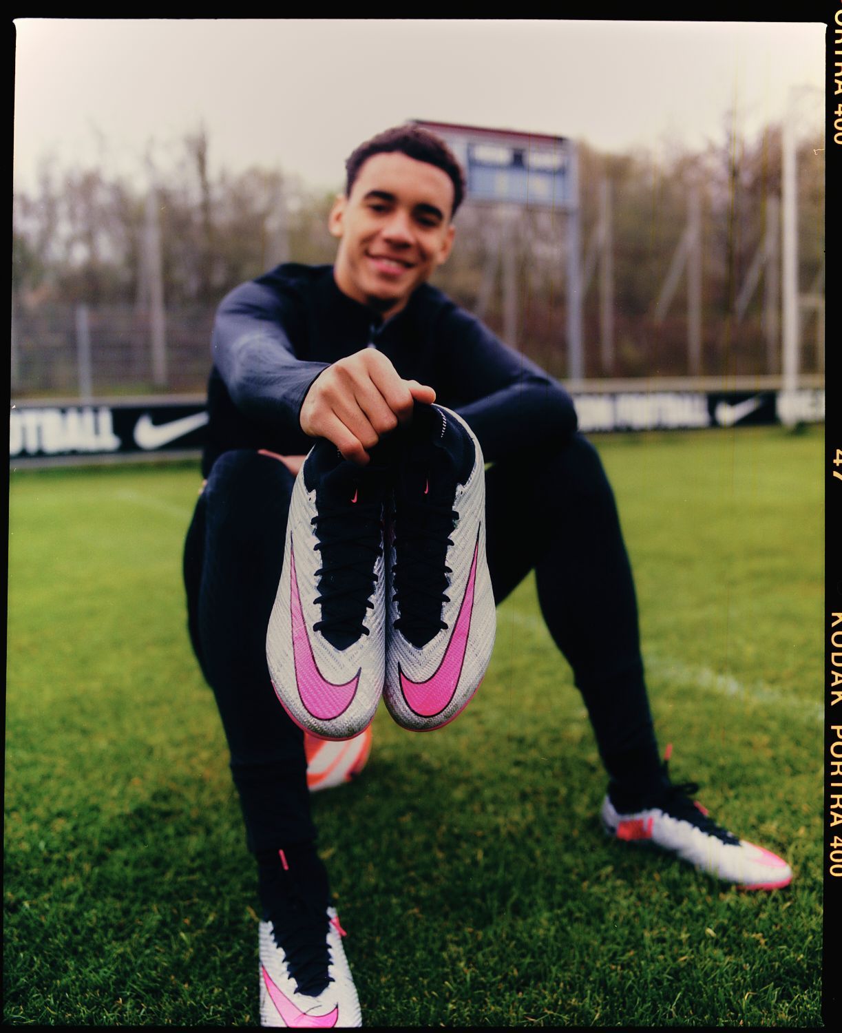 Lächelnder Fußballspieler, der auf einem Feld rosa und weiße Stollenschuhe hochhält.