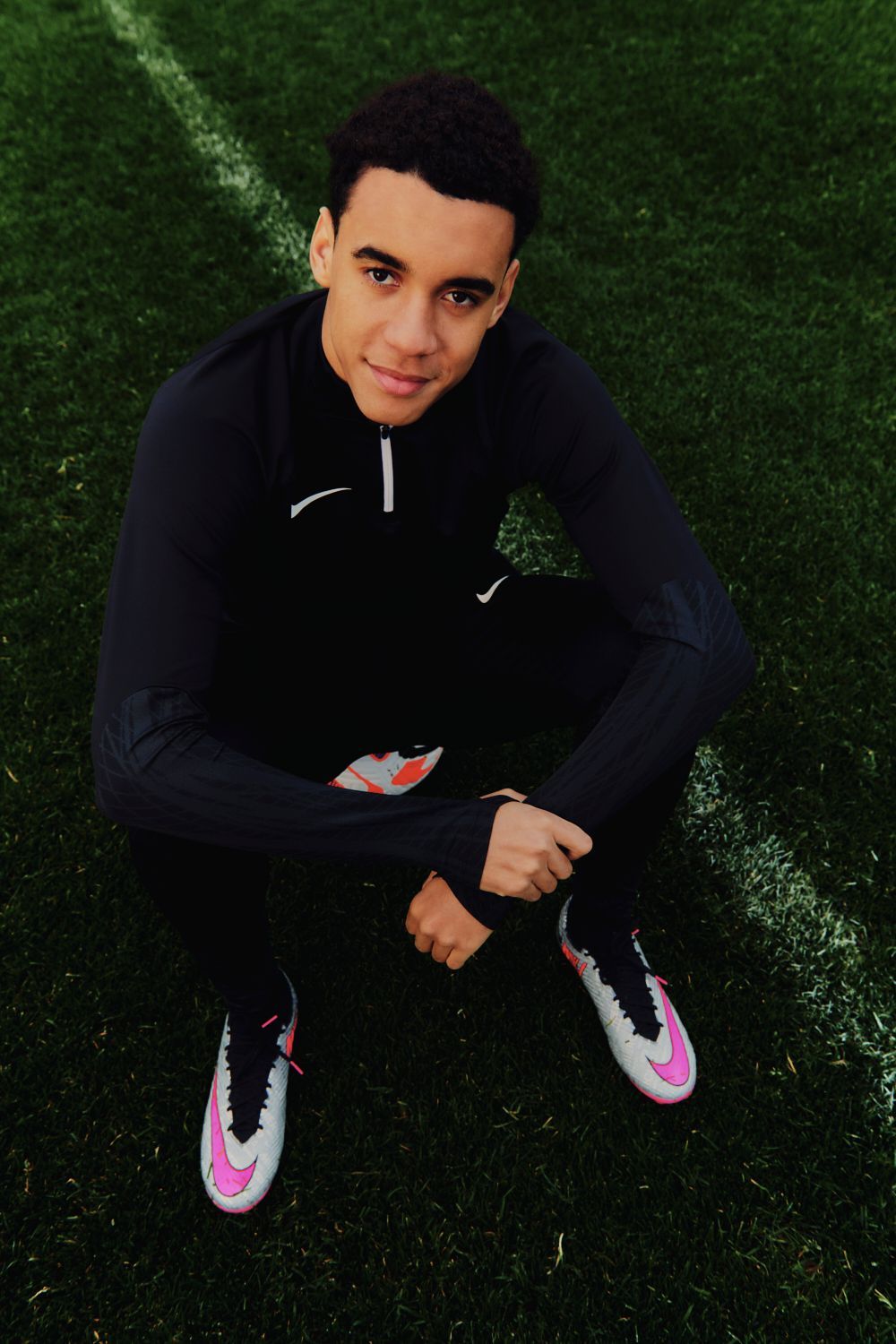 Junger Mann sitzt auf einem Fußballfeld, trägt schwarze Trainingskleidung und lächelt.