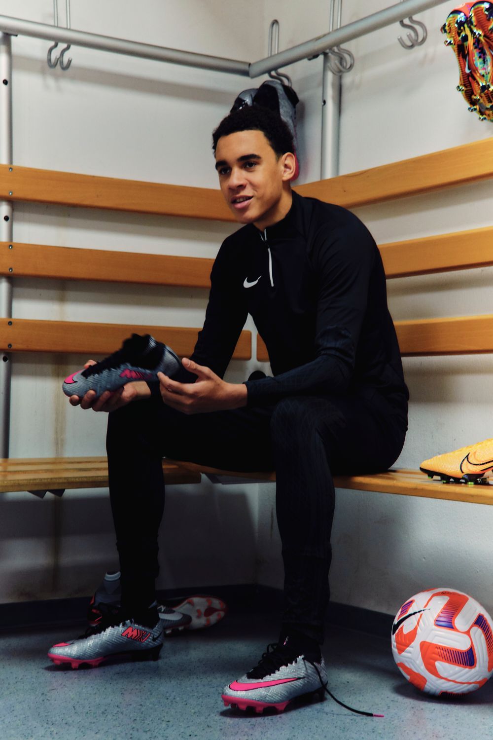 Mann sitzt mit Fußballschuhen in der Hand in der Umkleidekabine und trägt schwarze Sportkleidung.