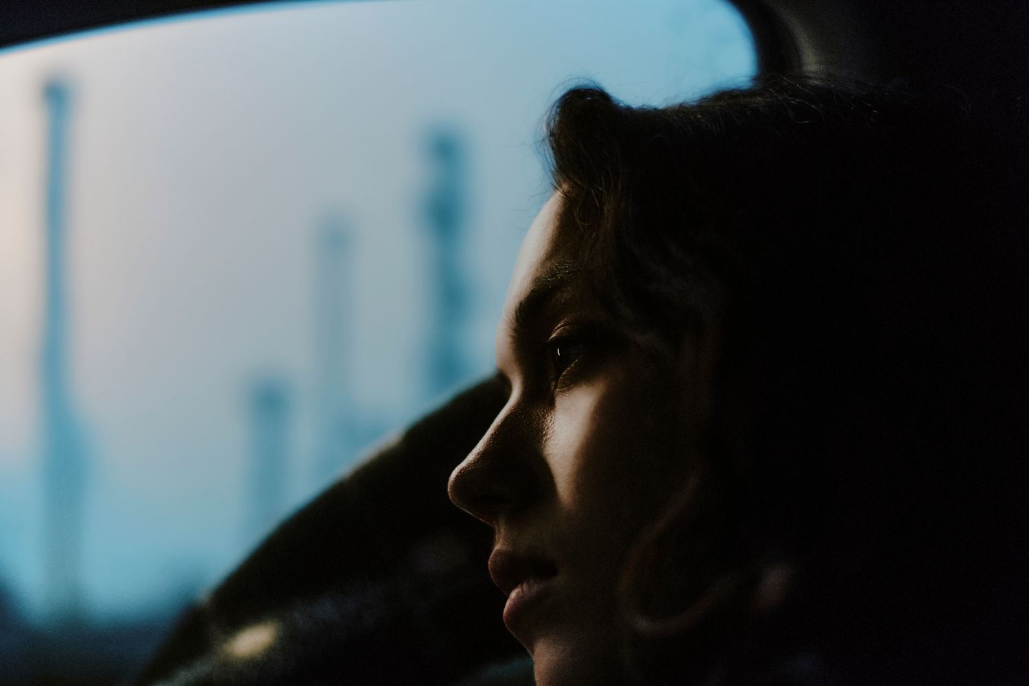 Frau blickt aus Autofenster auf Industrielandschaft; kühle Töne, nachdenklicher Ausdruck.