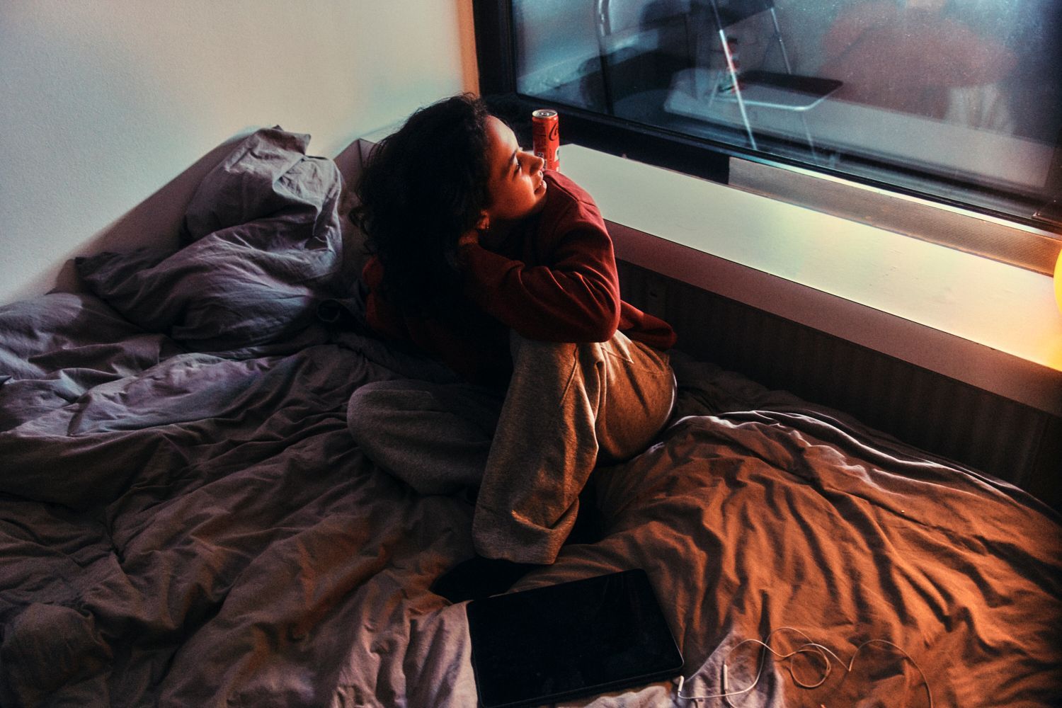 Frau sitzt auf dem Bett und schaut aus dem Fenster. Das Schlafzimmer ist mit warmem Licht beleuchtet, draußen ist es bewölkt.