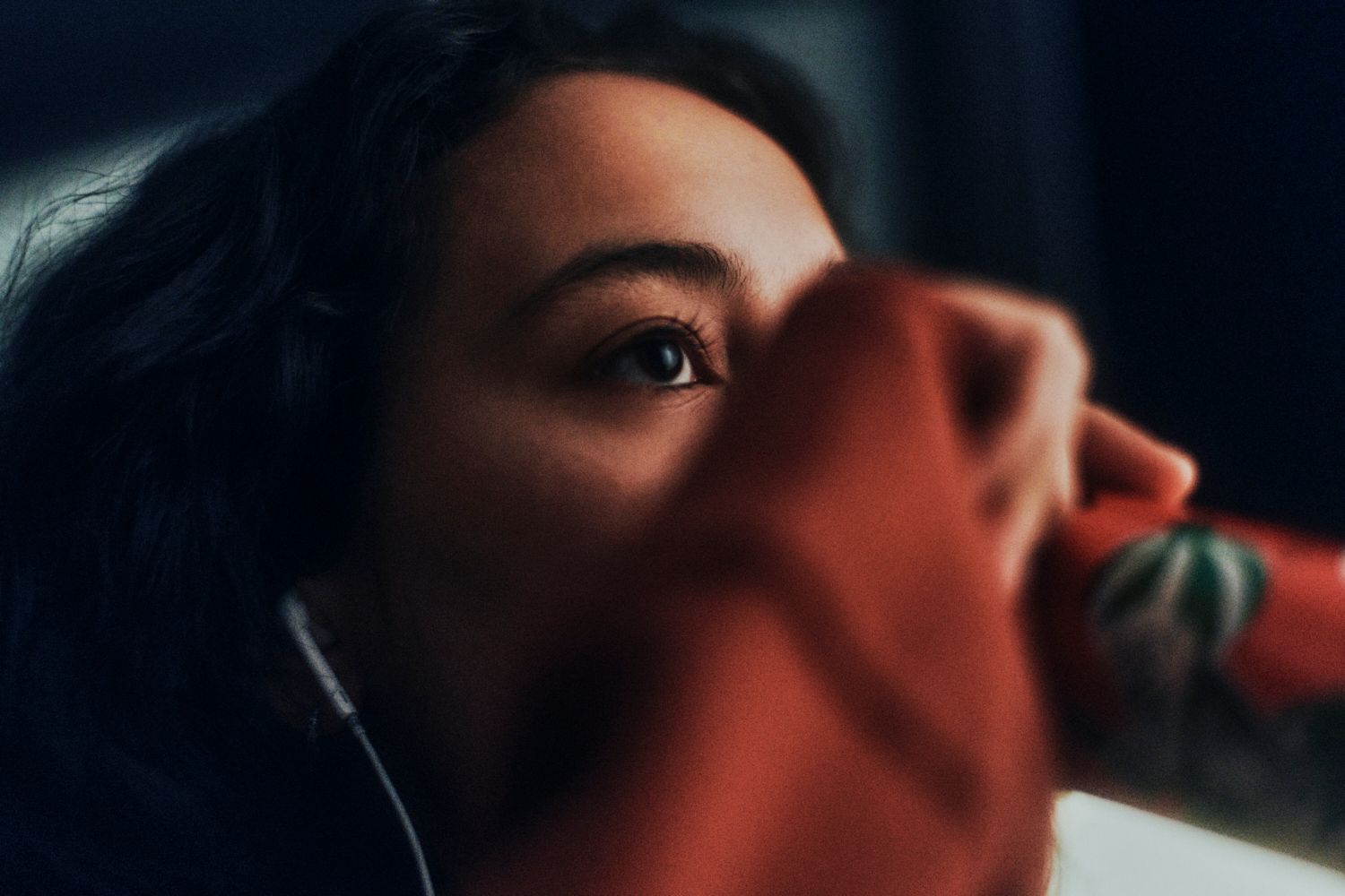 Frau mit dunklem Haar, nach oben schauend, eine rote Tasse mit Kaktusmuster in der Hand, Ohrhörer drin.