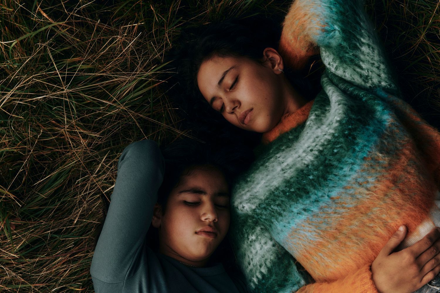 Zwei junge Menschen liegen schlafend im hohen Gras, der eine trägt einen bunten Pullover, der andere ein blaues Hemd.
