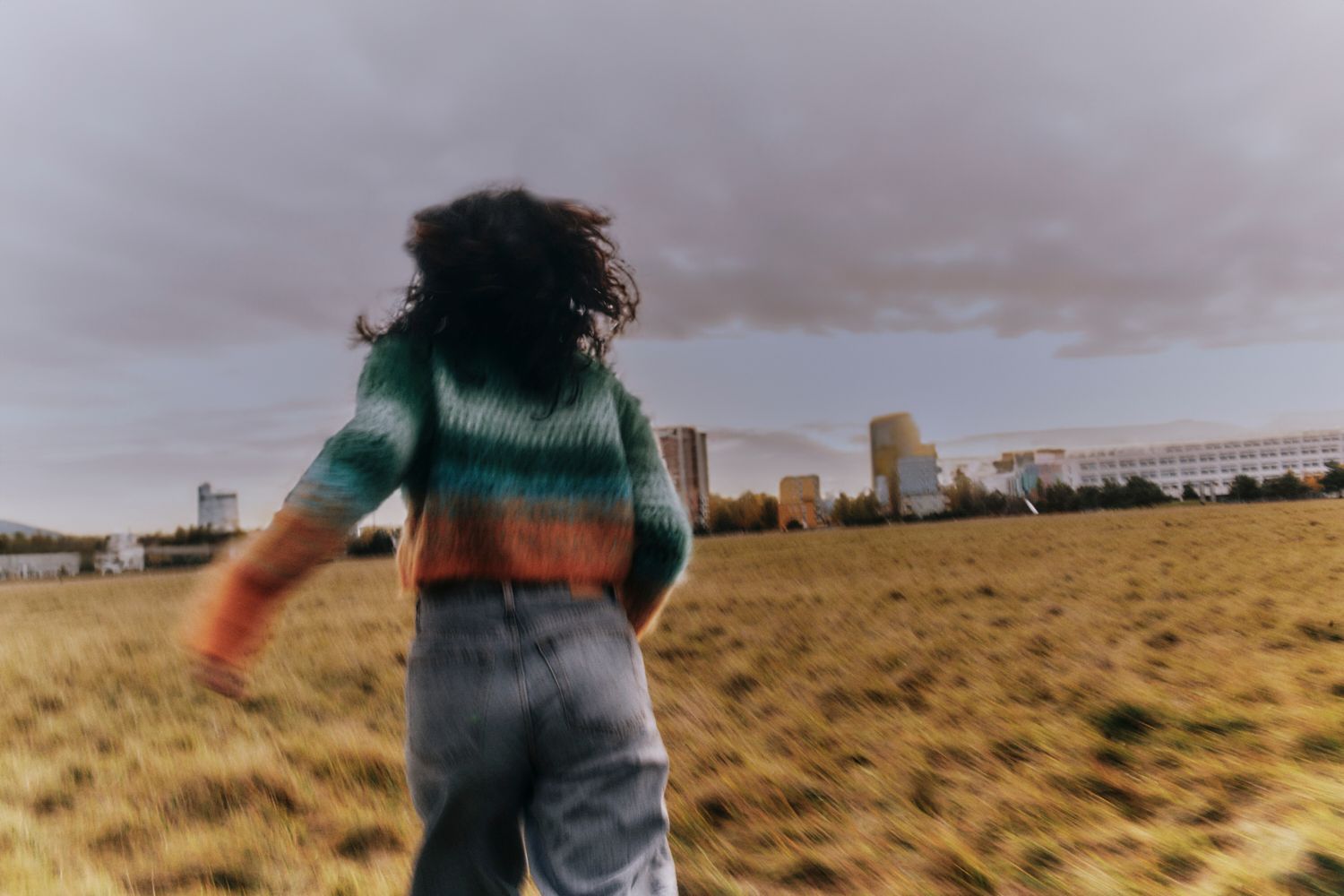 Person, die auf einem Feld mit Gebäuden im Hintergrund läuft; trägt einen gestreiften Pullover und Jeans.