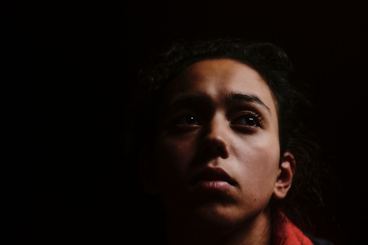 Frau blickt nach oben, beleuchtetes Gesicht vor schwarzem Hintergrund, trauriger Gesichtsausdruck.