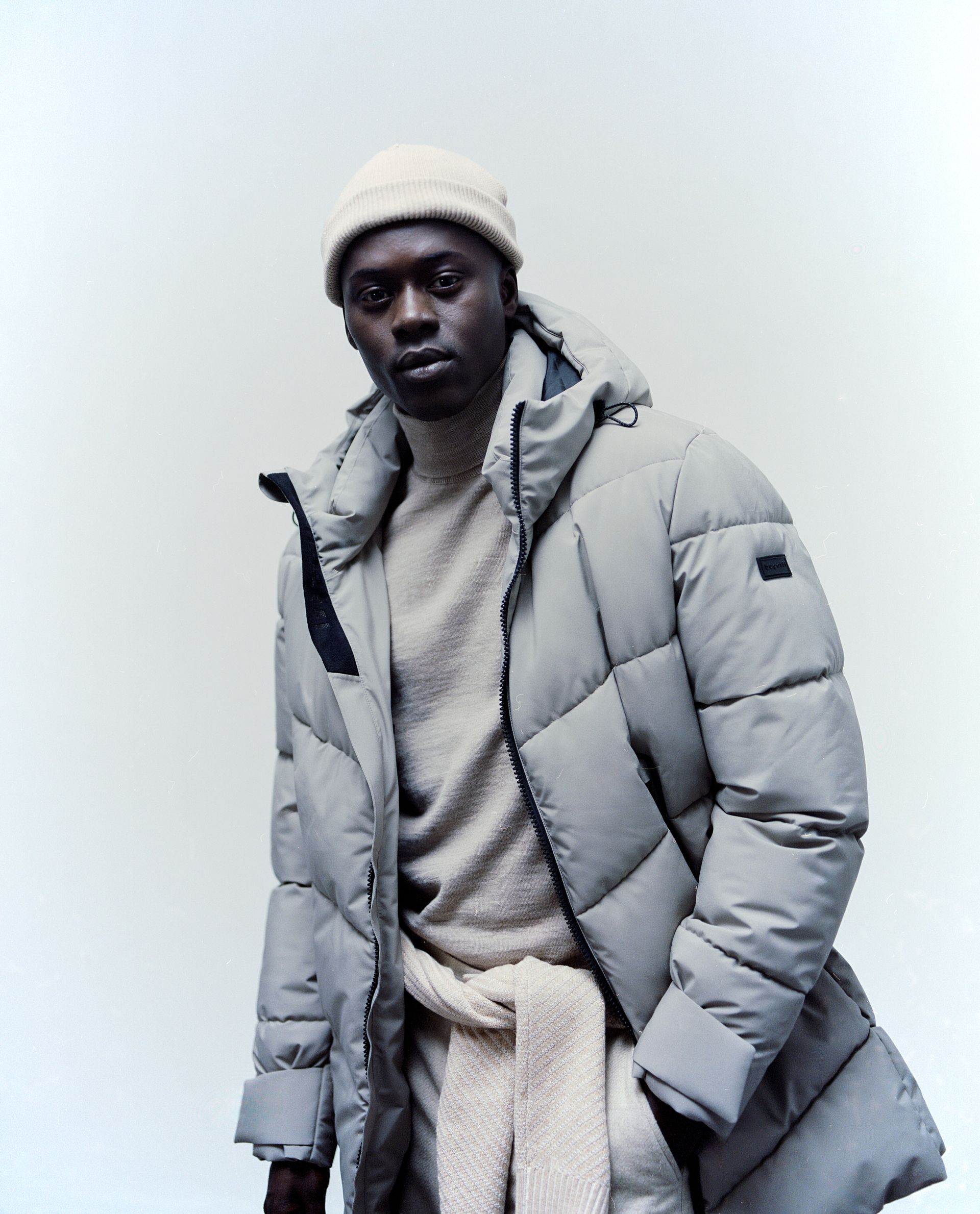 Mann mit hellgrauem Puffermantel, Beanie und Pullover vor neutralem Hintergrund.