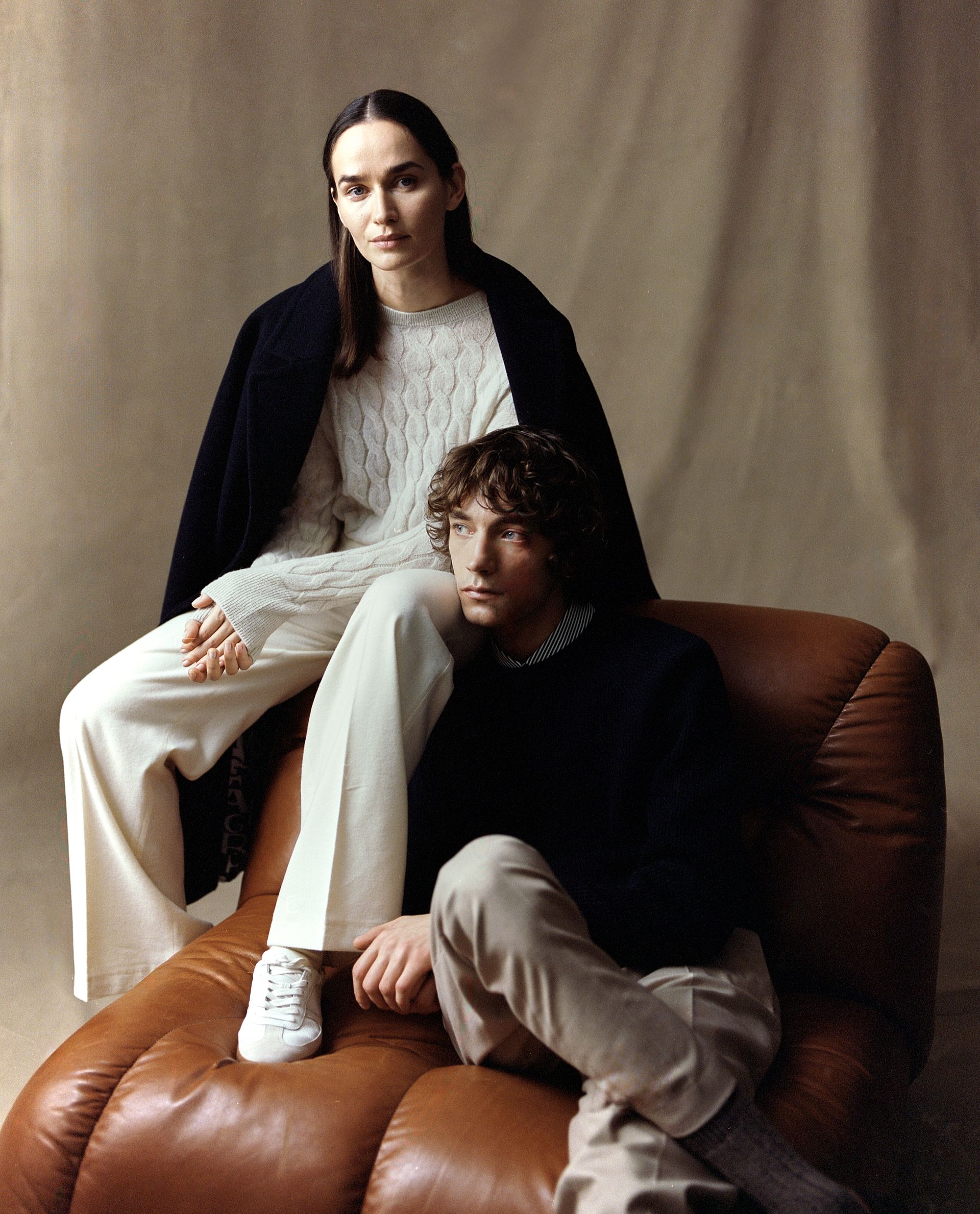 Zwei Personen sitzen auf einem braunen Ledersessel. Eine Frau in weißer Hose und Pullover sowie dunkler Jacke.