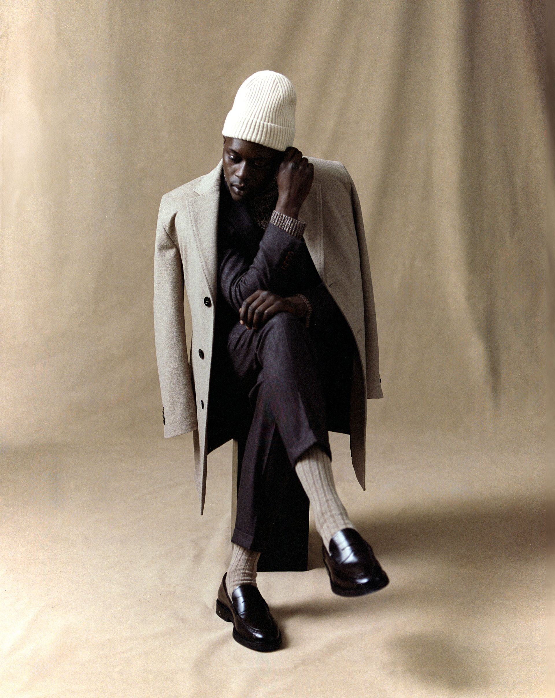 Mann mit weißer Mütze und Mantel, sitzt mit überkreuzten Beinen da und sieht nachdenklich aus.