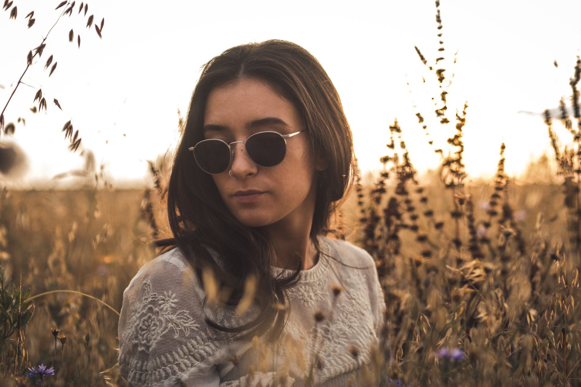Frau mit Sonnenbrille in einem Feld mit hohem Gras, goldenes Sonnenlicht, Blick nach unten.
