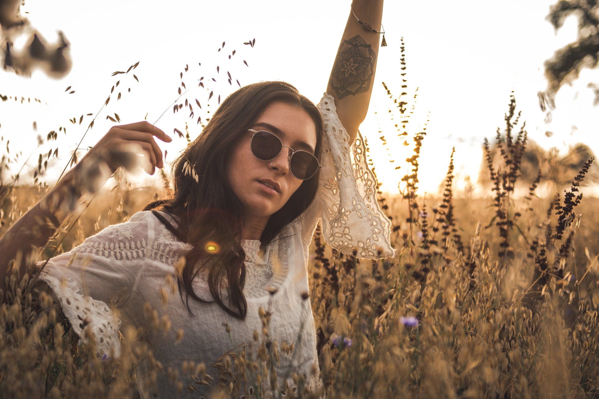 Frau mit Sonnenbrille und weißem Spitzentop posiert mit erhobenem Arm auf einem sonnenbeschienenen Feld.