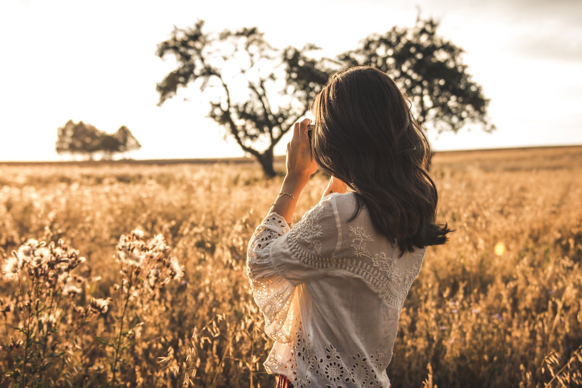 Frau in weißem Spitzenhemd fotografiert ein Feld bei Sonnenuntergang.