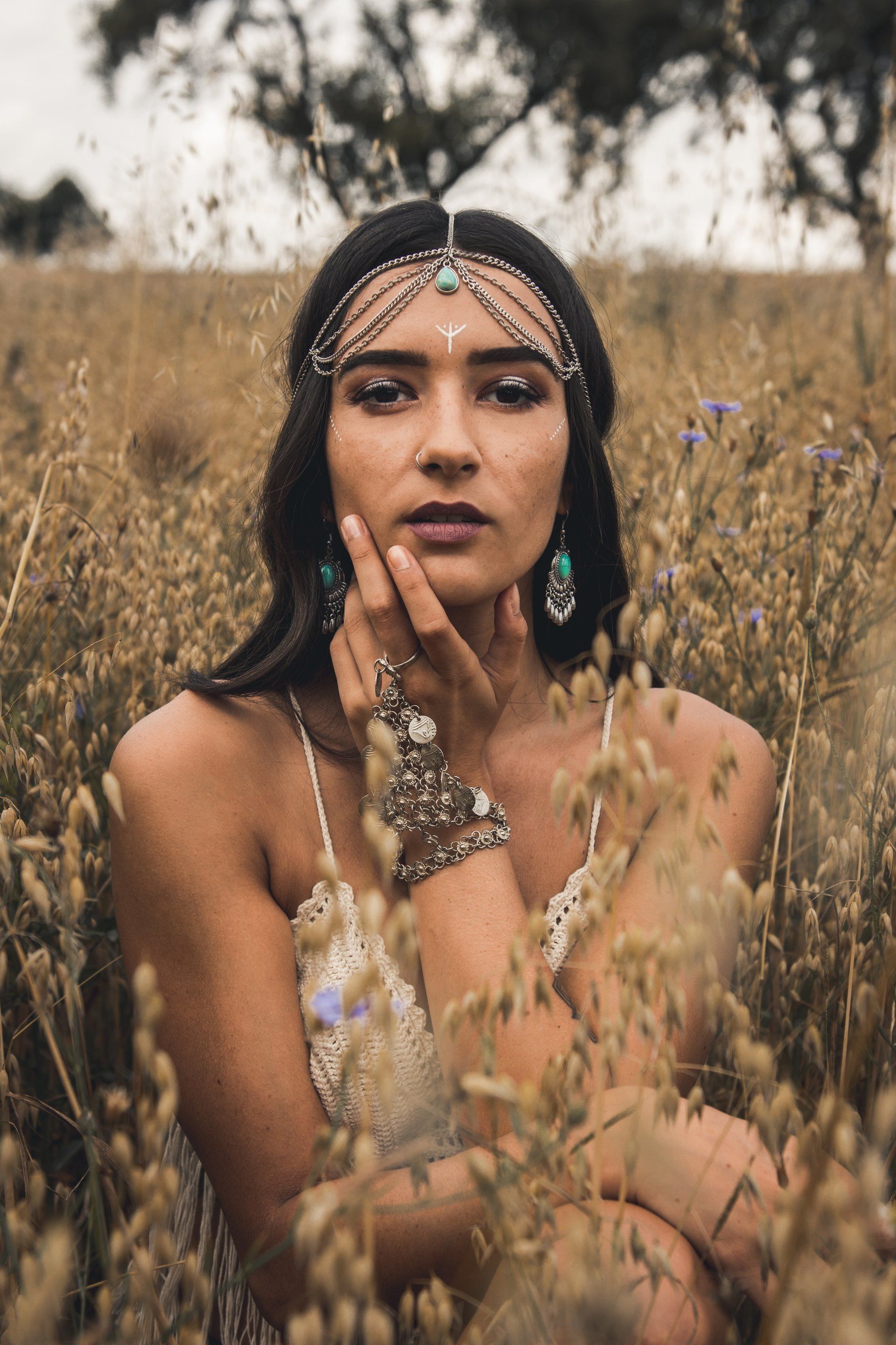 Frau auf einem Feld, trägt kunstvollen Schmuck, die Hand auf dem Gesicht, blickt direkt in die Kamera.