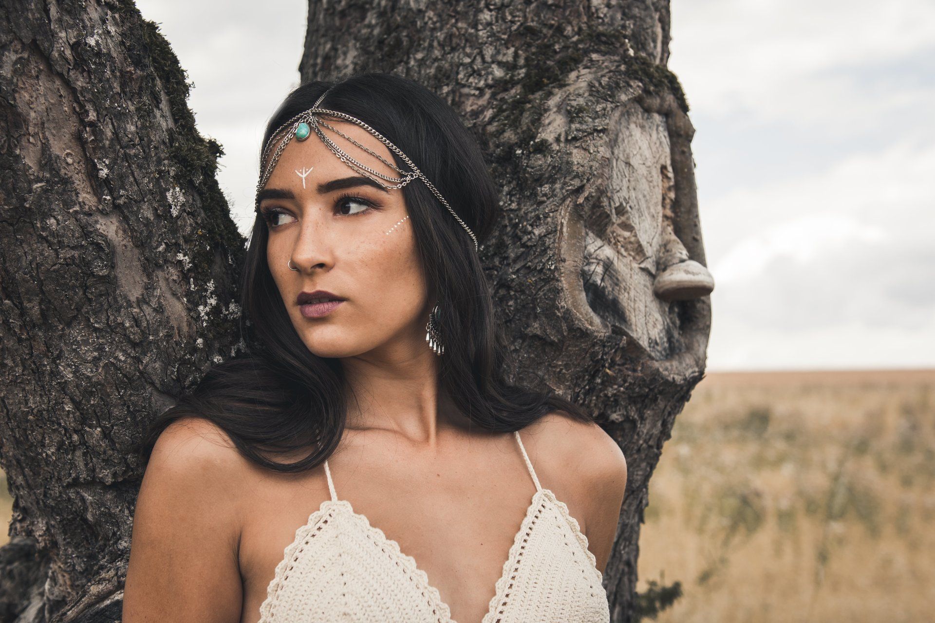 Frau mit Kopfbedeckung, an einem Baum auf einem Feld gelehnt; trägt ein Häkeloberteil und schaut weg.