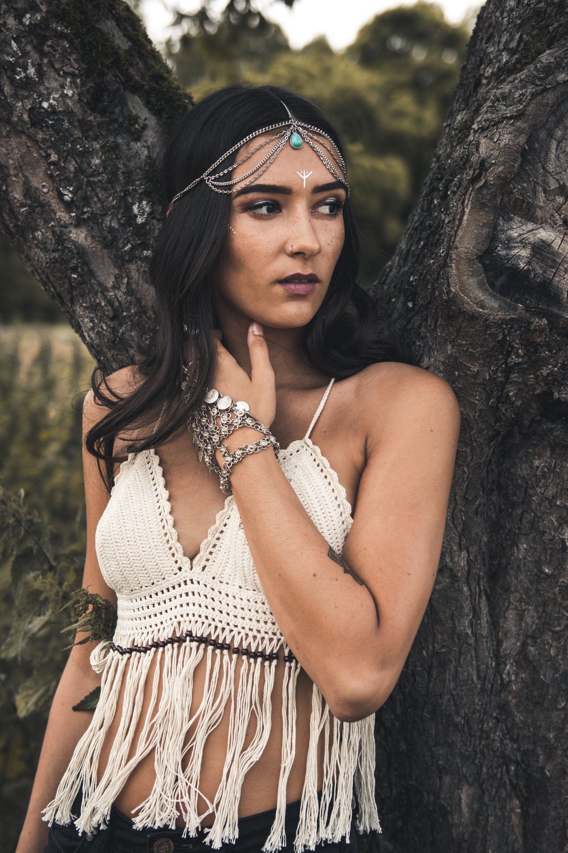 Frau mit gehäkeltem bauchfreiem Top und Kopfschmuck, an einem Baum gelehnt, Blick zur Seite gerichtet.
