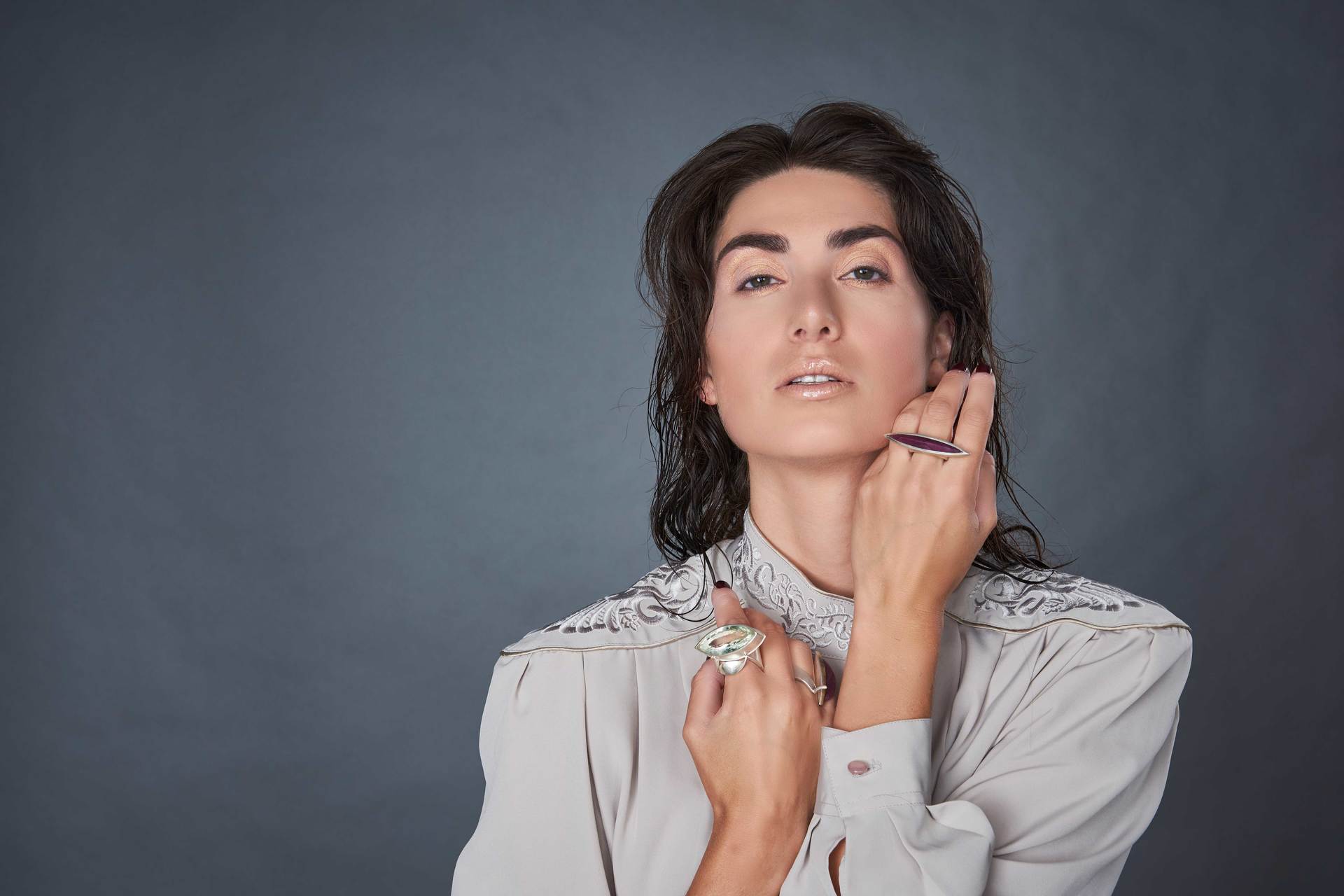 Frau mit nassen Haaren und Ringen, trägt ein helles Hemd, die Hände erhoben, vor einem grauen Hintergrund.