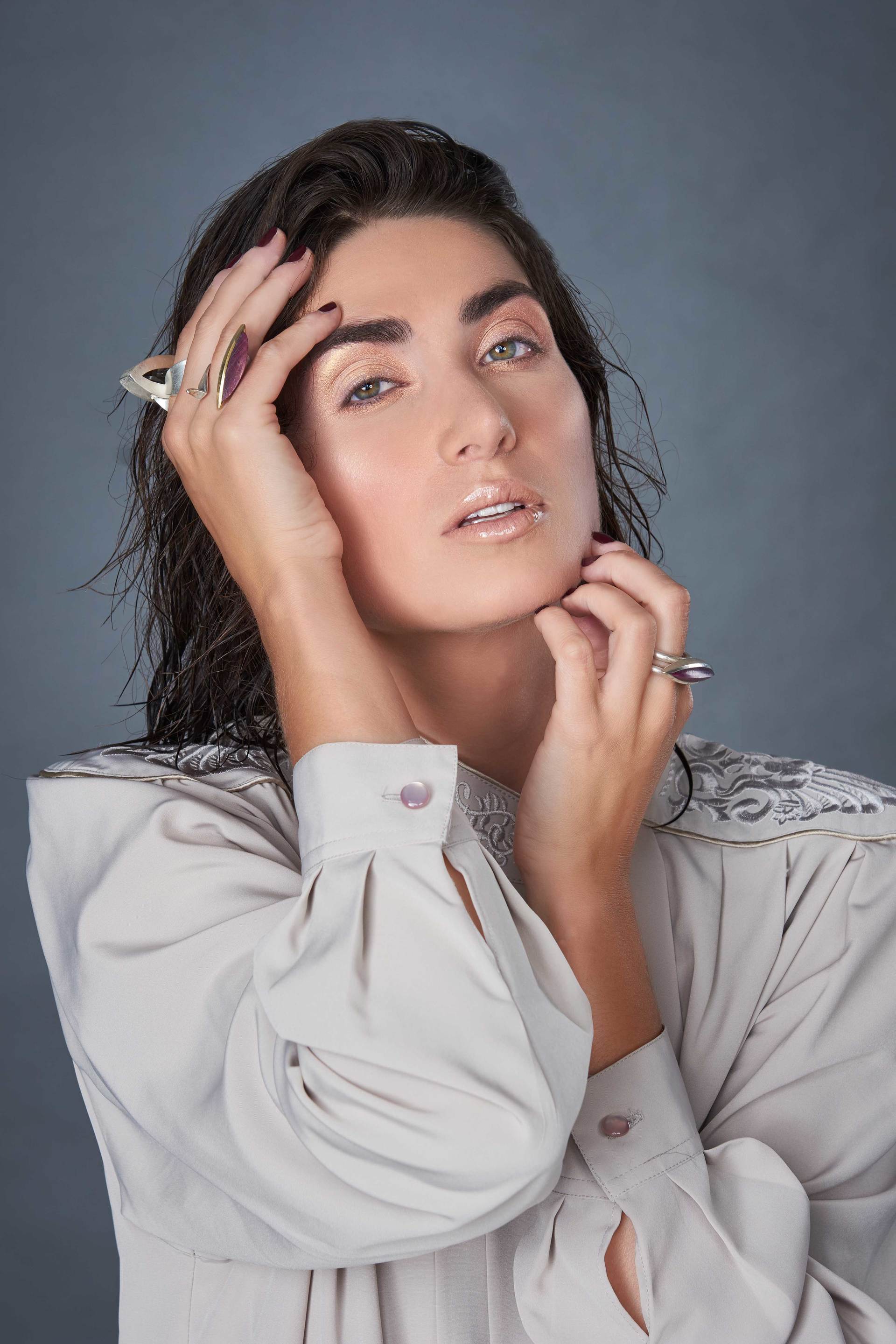 Frau mit dunklem Haar, leichtem Make-up, trägt eine helle Bluse und vergräbt die Hände im Gesicht.