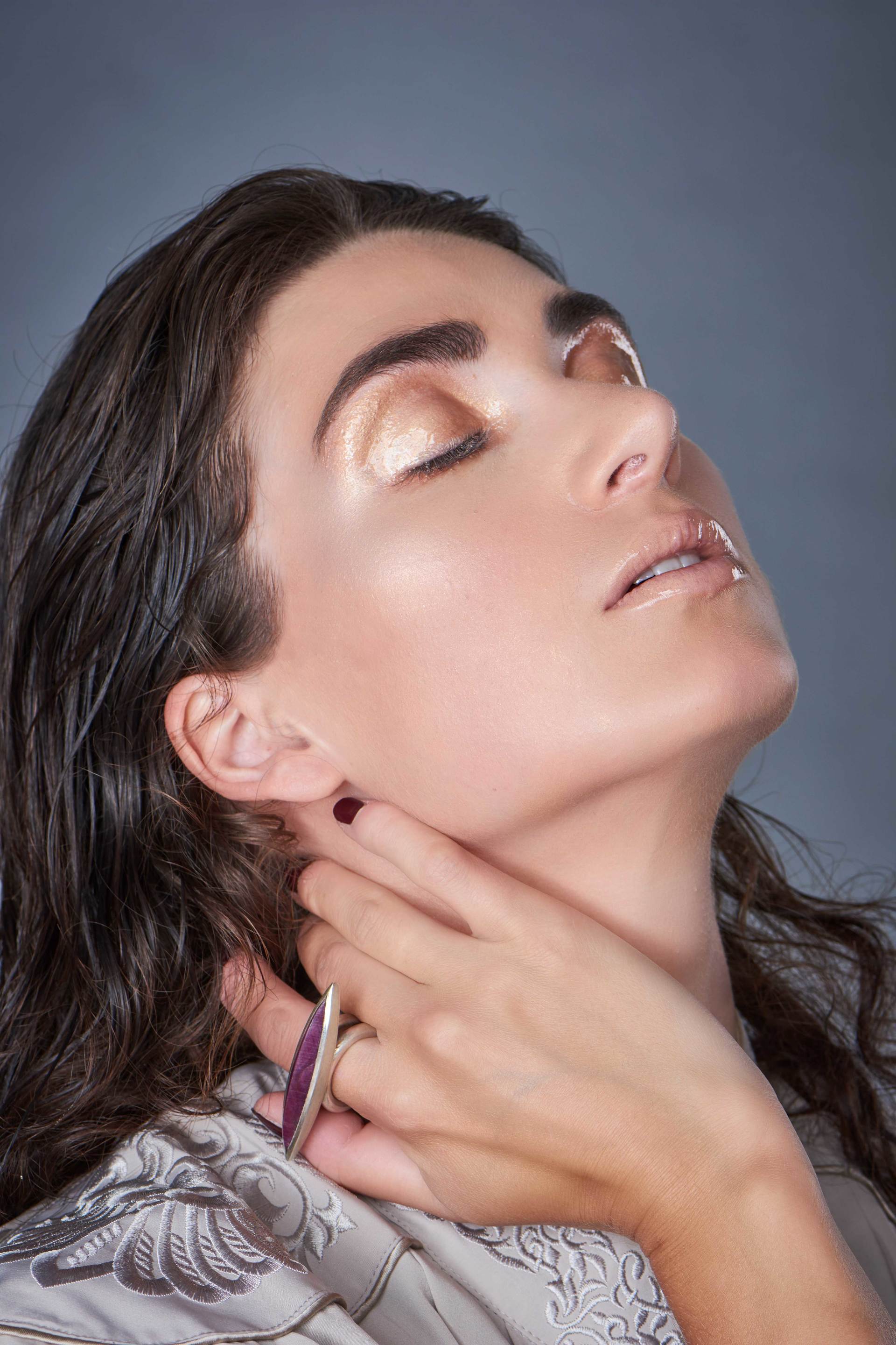 Frau mit geschlossenen Augen, Hand am Hals, geschminkt und mit Ring, vor grauem Hintergrund.
