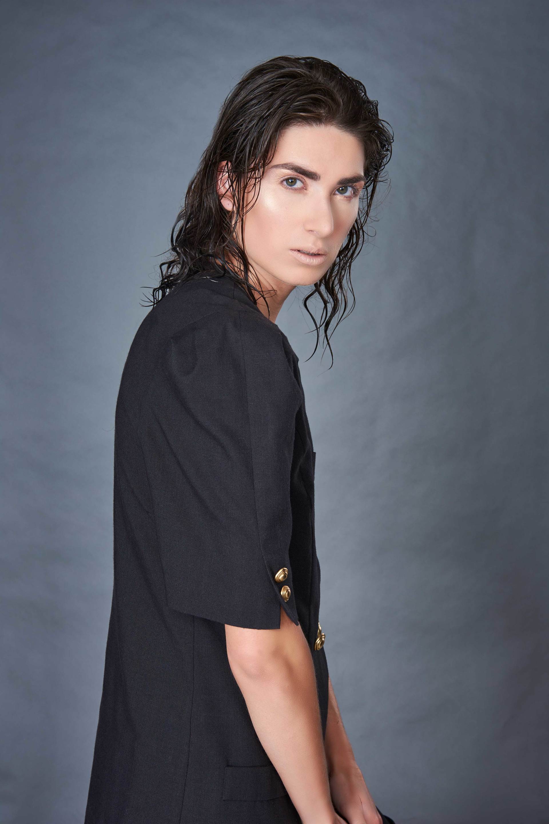 Eine Person mit dunklem, nassem Haar in einer schwarzen Jacke, die über die Schulter blickt, vor einem grauen Hintergrund.