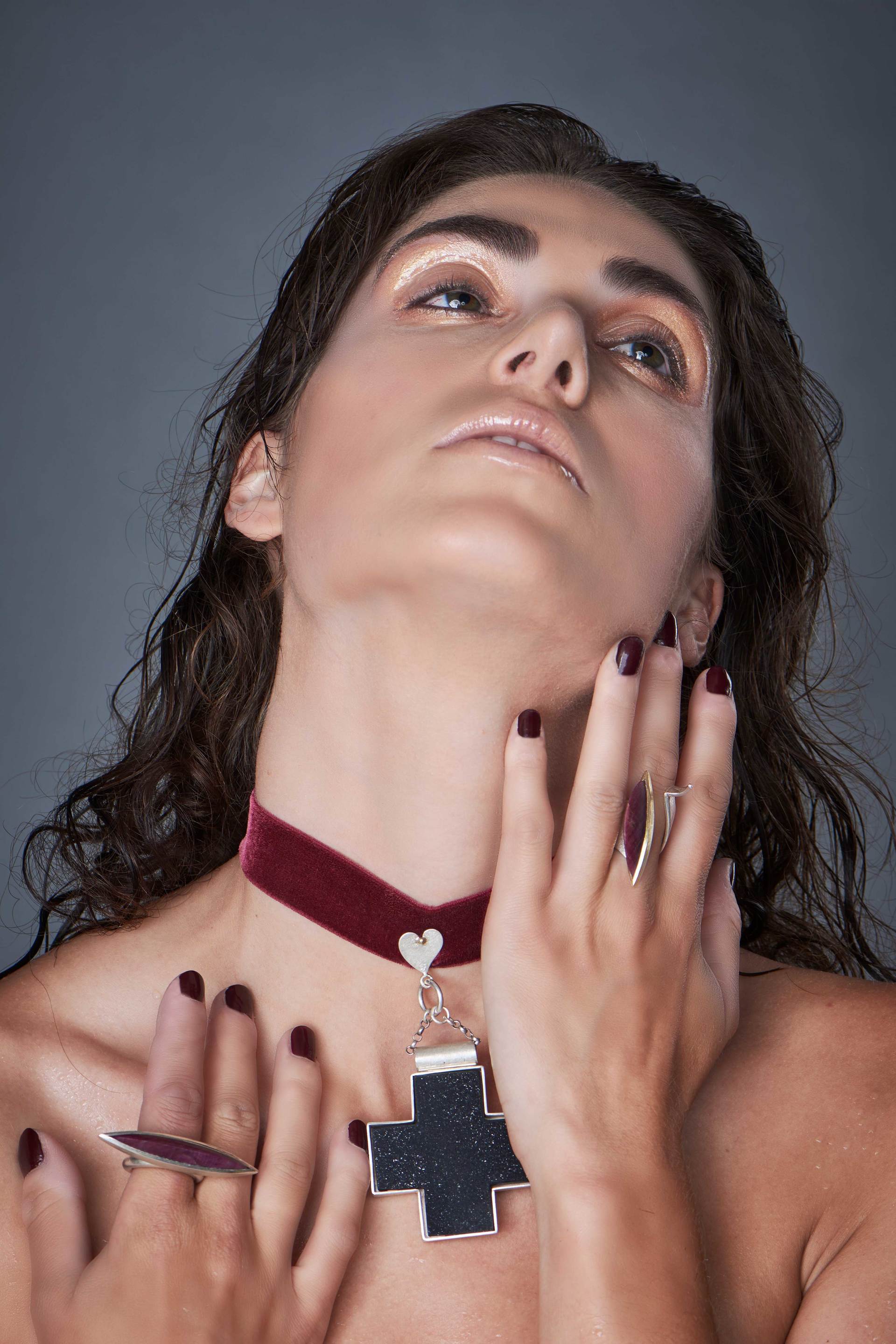 Frau mit dunklem Haar, einem roten Halsband und einer Kreuzkette sowie dunklem Nagellack, die nach oben blickt.