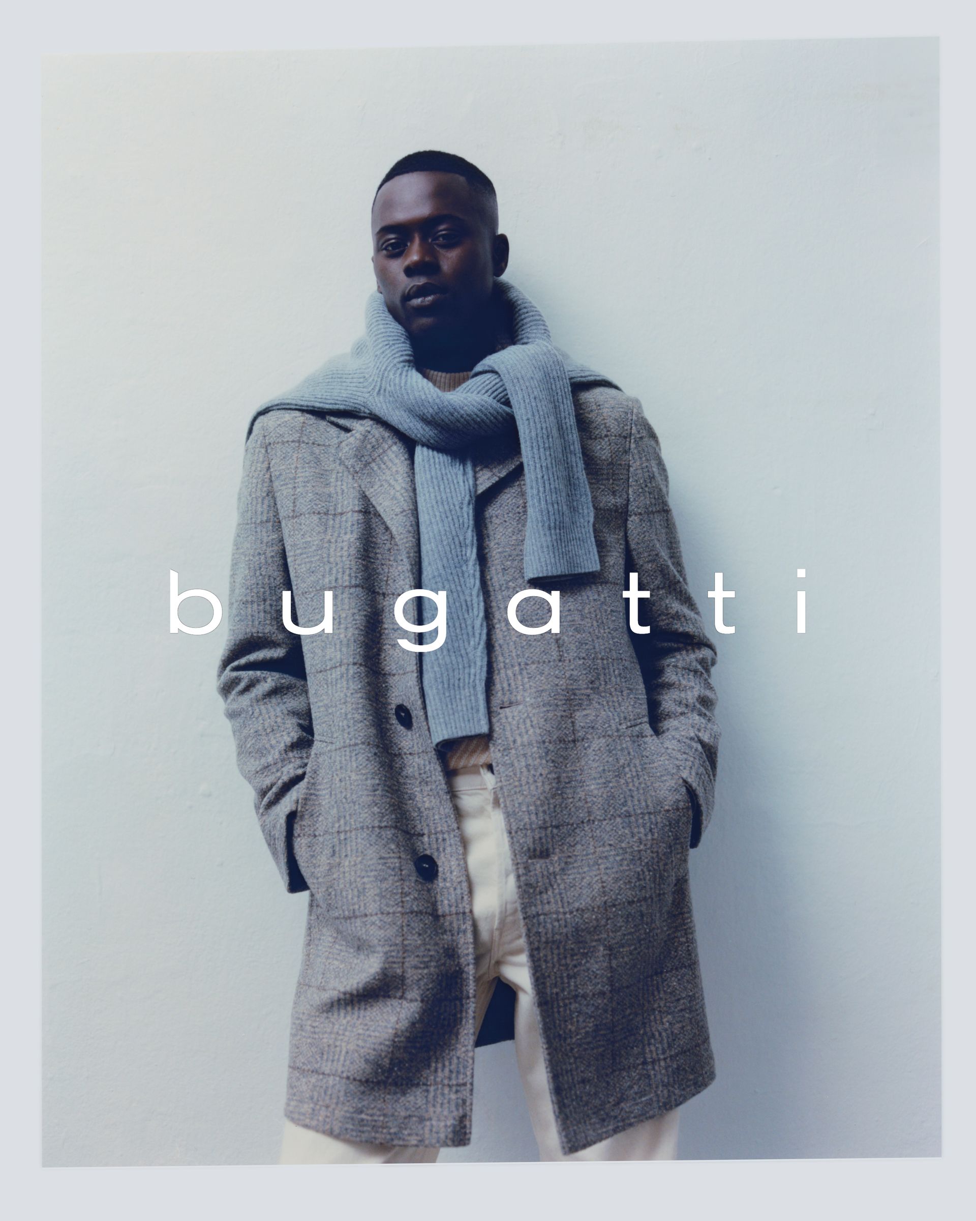 Mann mit grauem Mantel, hellblauem Schal und cremefarbener Hose posiert vor einer weißen Wand; Aufschrift „Bugatti“.