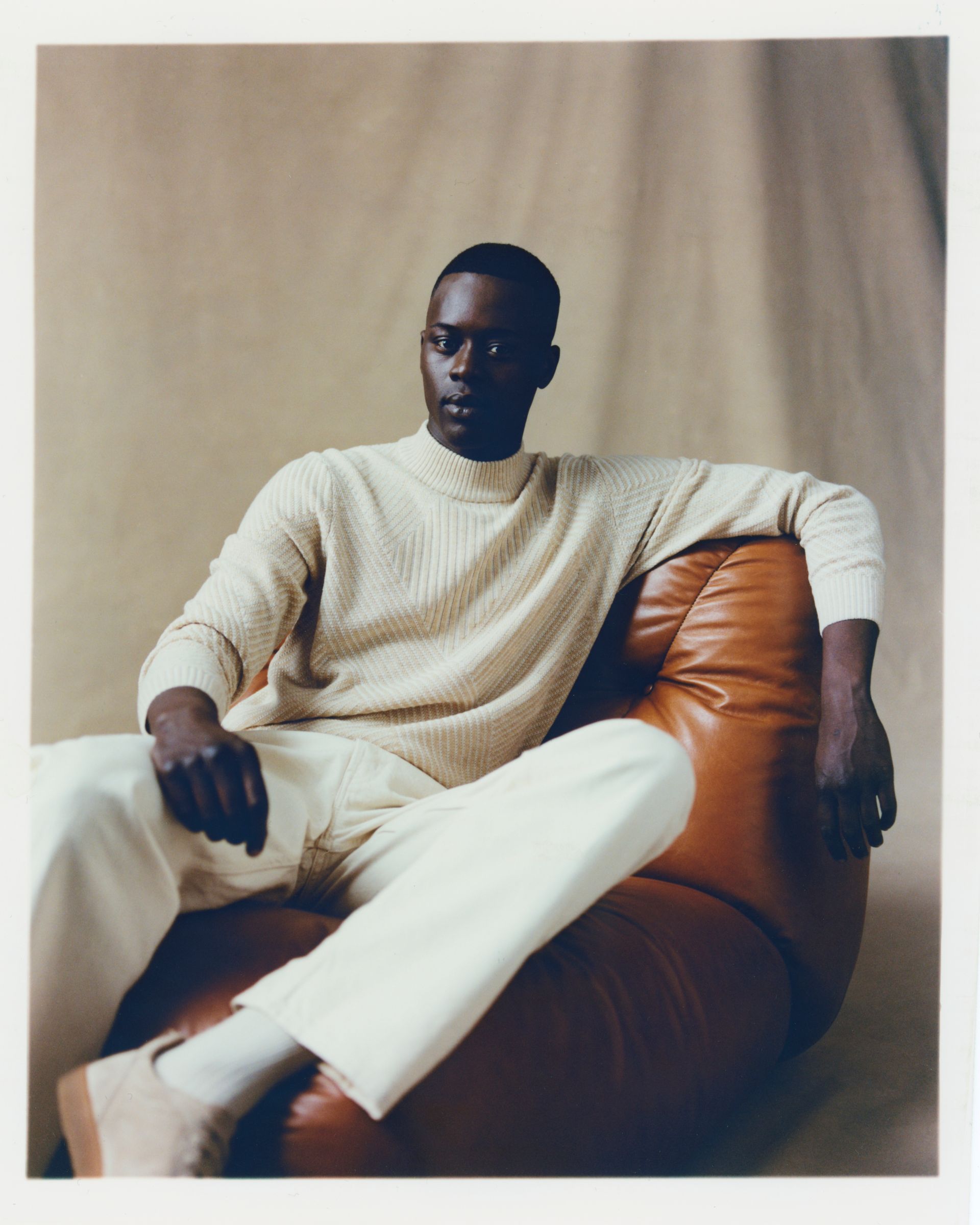 Mann in cremefarbenem Pullover und Hose, sitzt in einem braunen Ledersitzsack und schaut zur Seite.