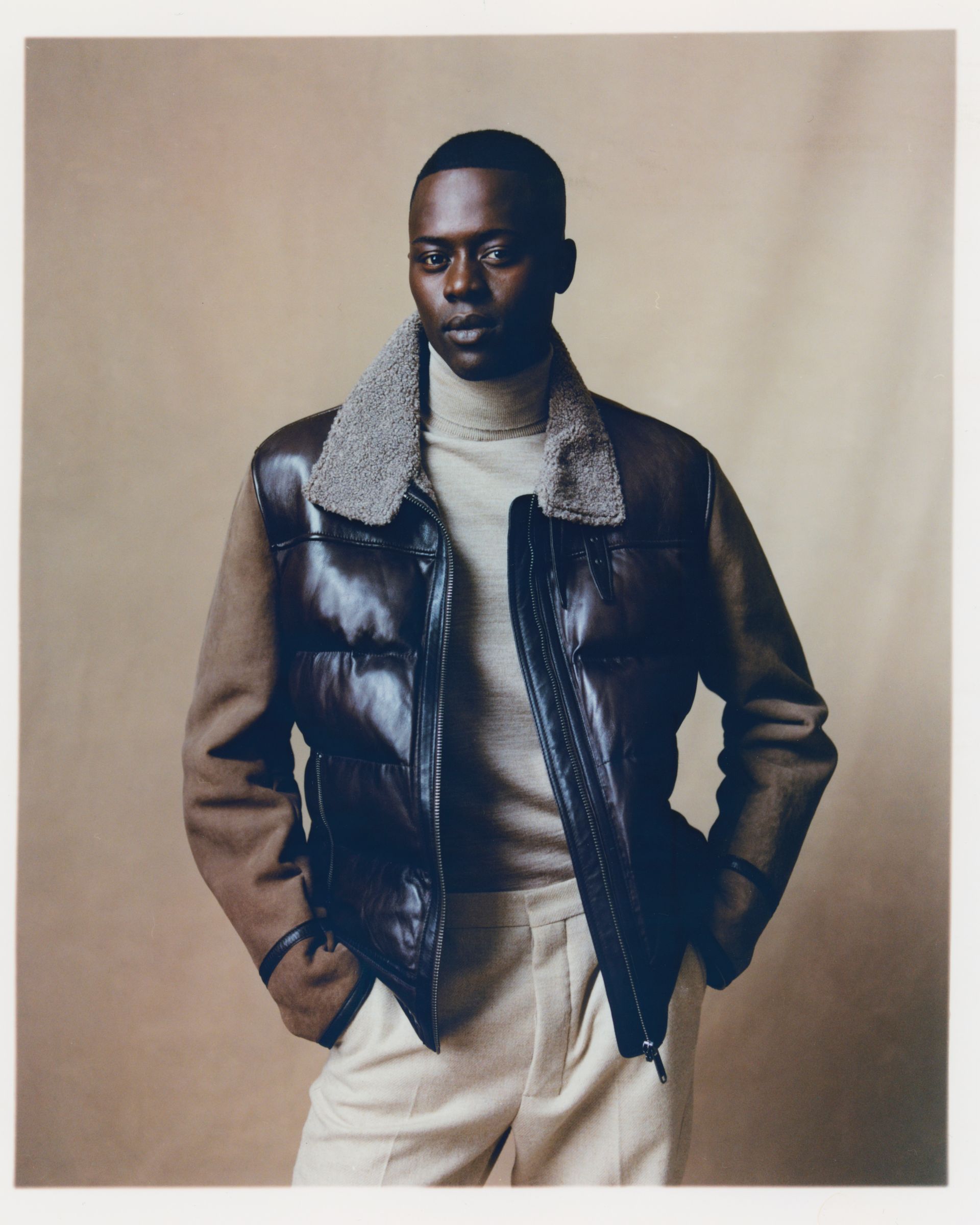 Mann in brauner Lederjacke, Rollkragenpullover und cremefarbener Hose, Hände in den Taschen, vor neutralem Hintergrund.