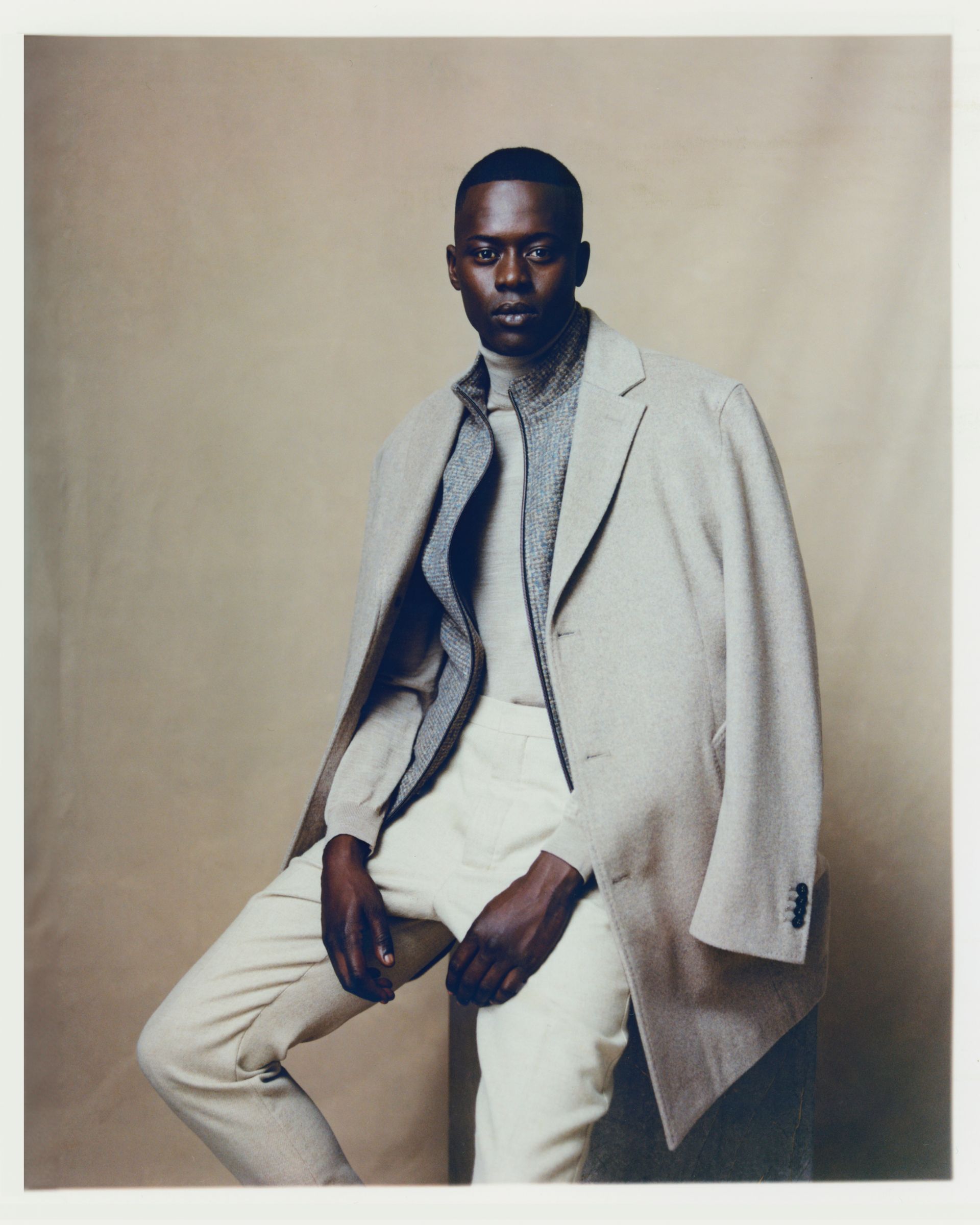 Ein Mann im beigen Anzug sitzt nach vorne gebeugt vor einem neutralen Hintergrund.