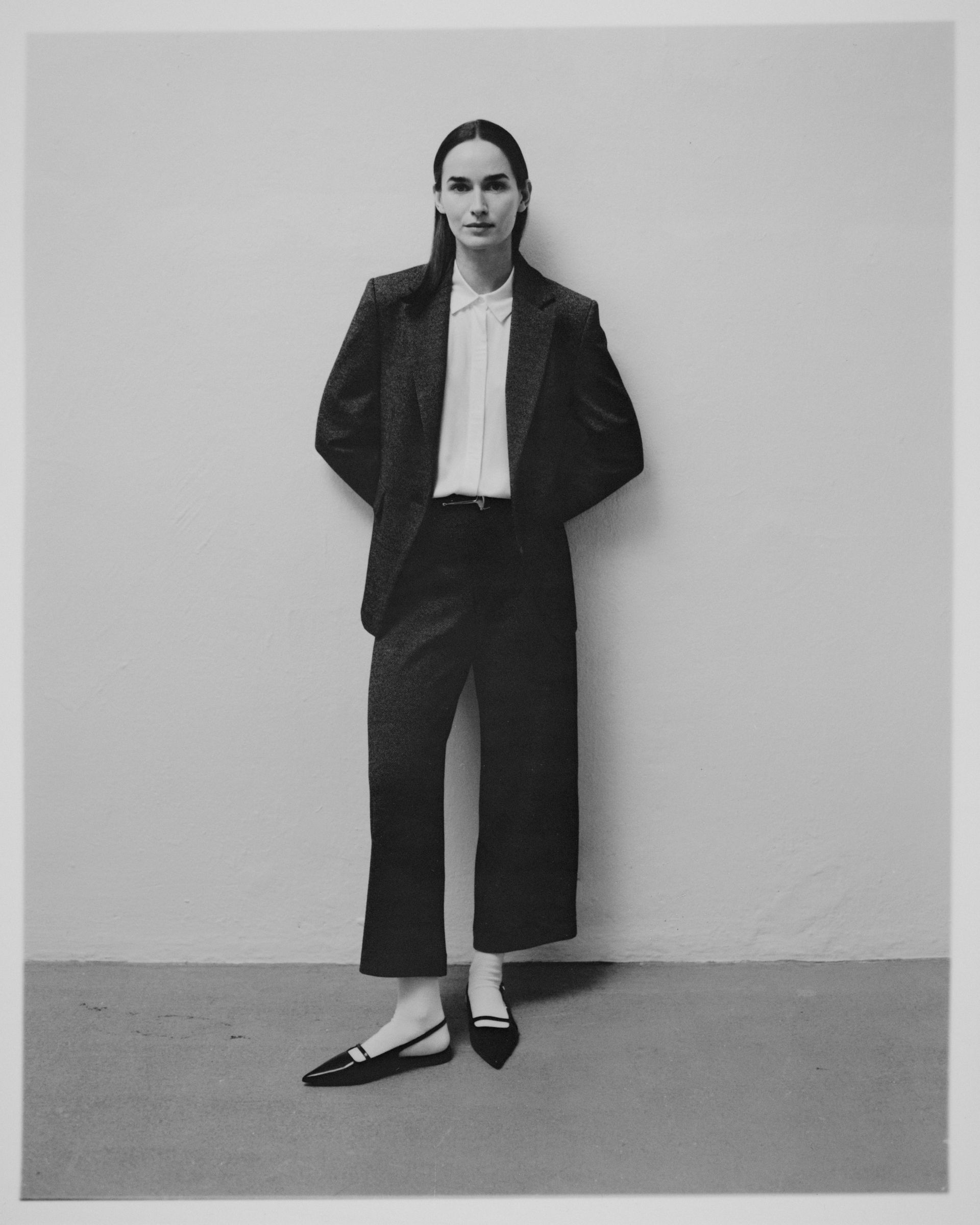 Frau in Blazer, Bluse und Hose, stehend mit den Händen hinter dem Rücken; einfarbig.