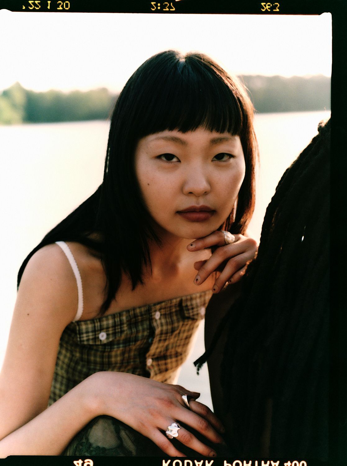 Frau mit dunklem Haar und Pony, trägt ein kariertes Oberteil und legt die Hand auf das Kinn.