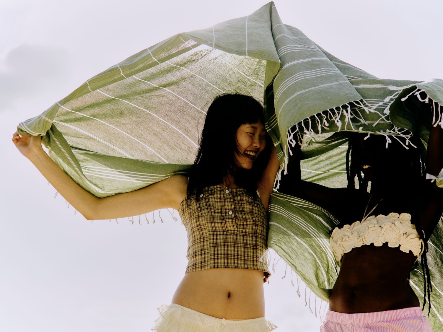 Zwei Frauen halten eine grün-weiß gestreifte Decke und lachen im hellen Sonnenlicht.