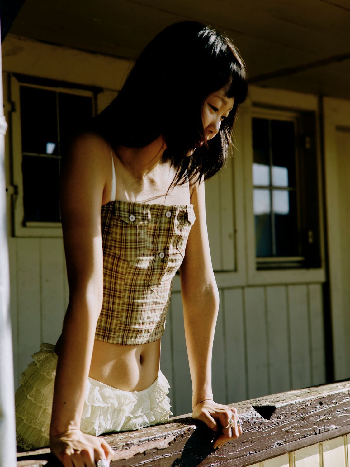 Frau in bauchfreiem Top und Rock lehnt an einem Holzgeländer und blickt nach unten. Sonnendurchfluteter Raum.