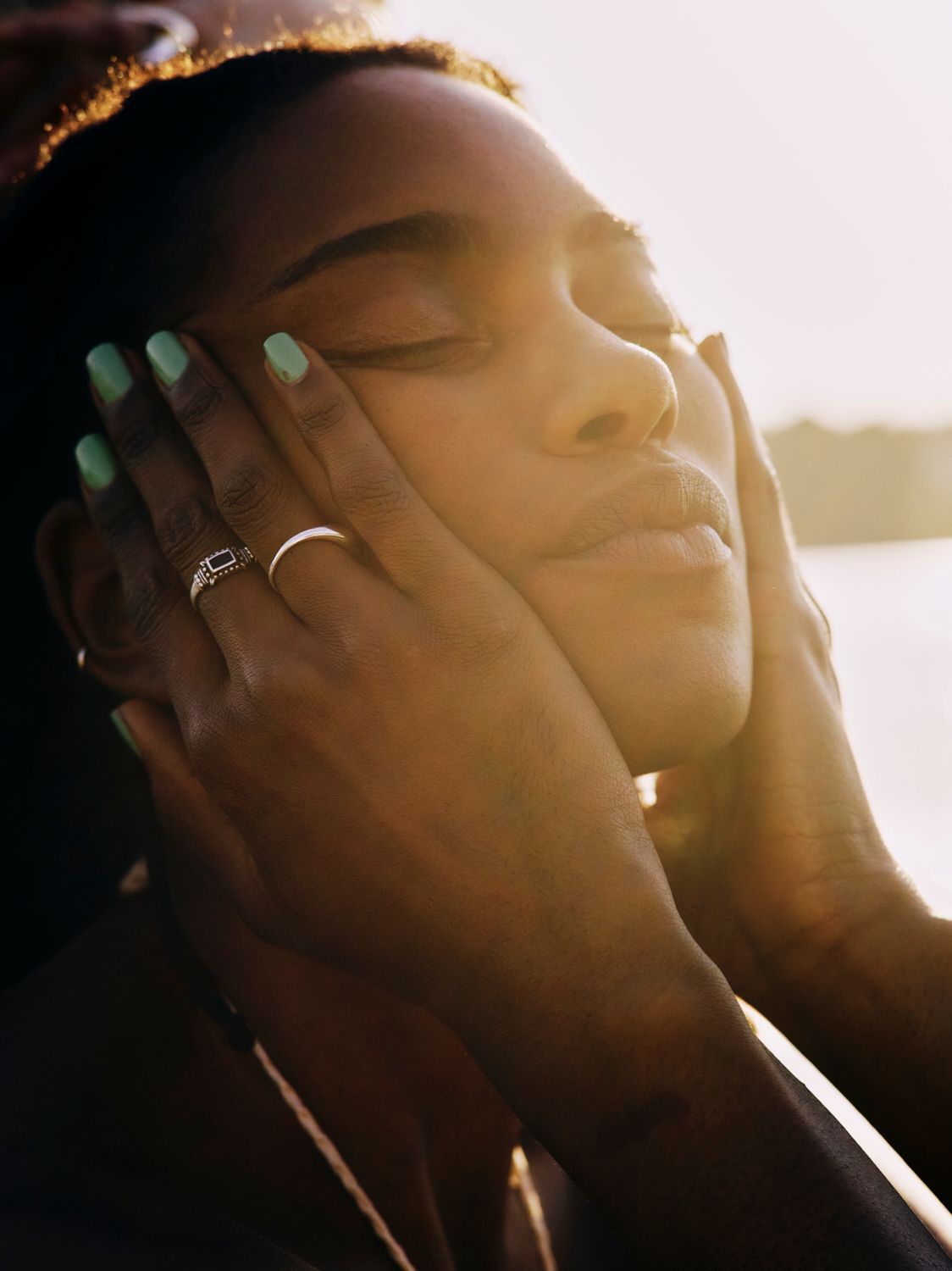 Frau mit geschlossenen Augen berührt ihr Gesicht mit den Händen; grüne Nägel. Goldenes Sonnenlicht.