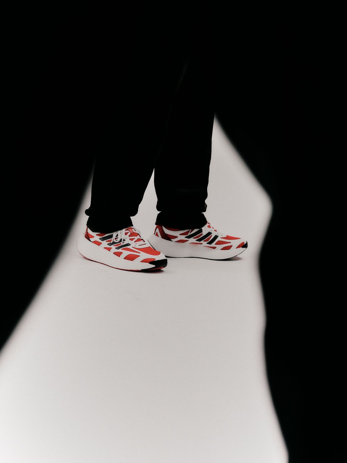 Person in schwarzer Hose und rot-weiß gemusterten Turnschuhen, die auf einer weißen Fläche steht.