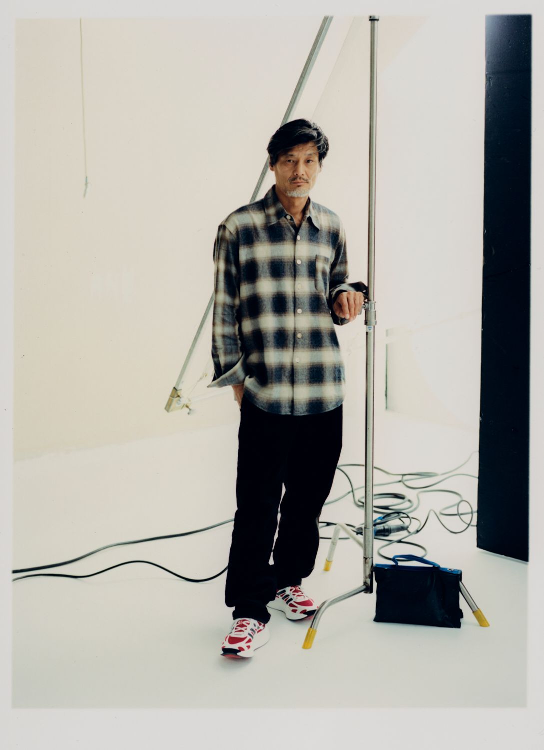 Ein Mann in kariertem Hemd und Turnschuhen steht in einem Studio und hält ein Lichtstativ.