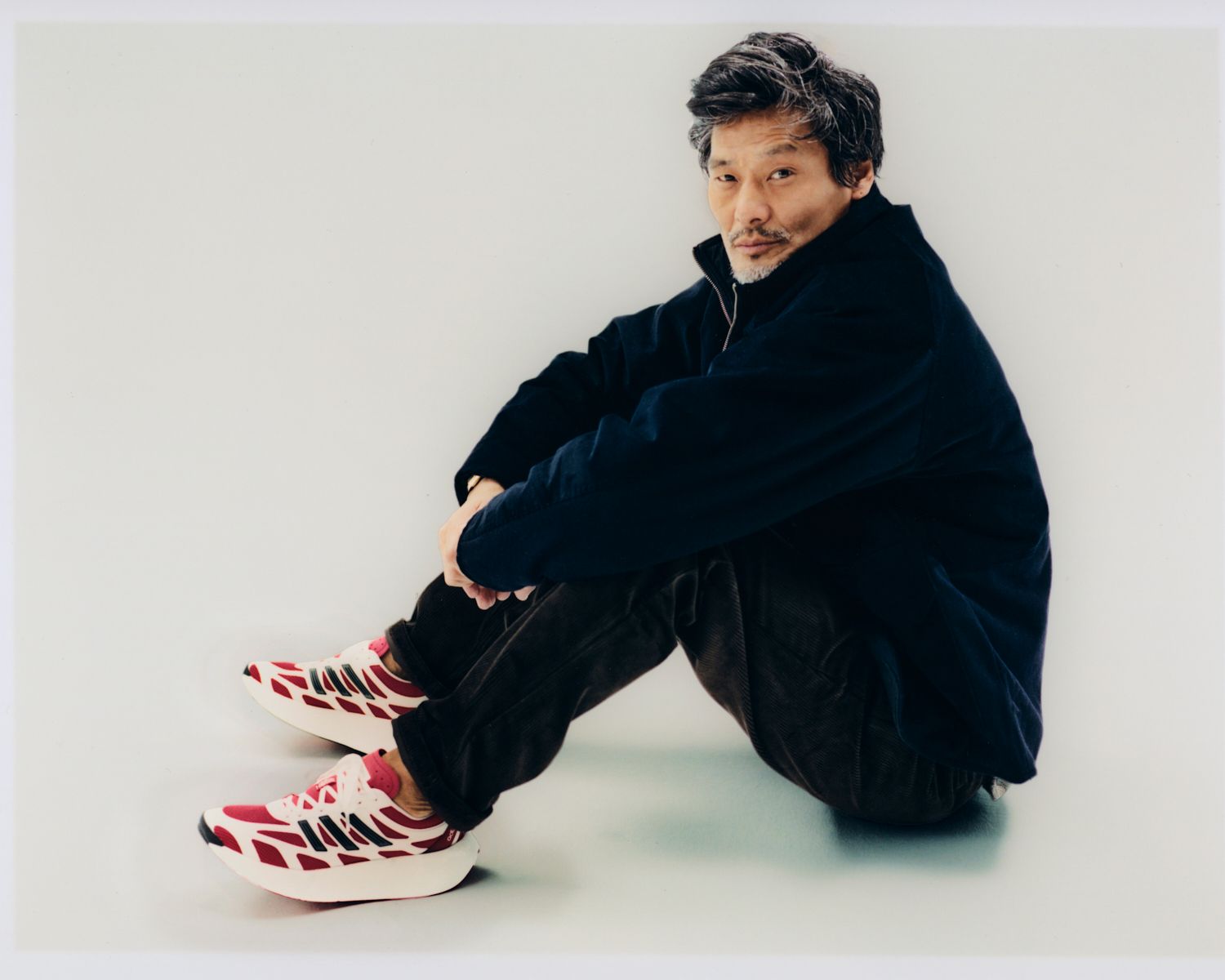 Mann sitzt mit überkreuzten Beinen, trägt eine marineblaue Jacke, braune Hosen und rot-weiße Turnschuhe.