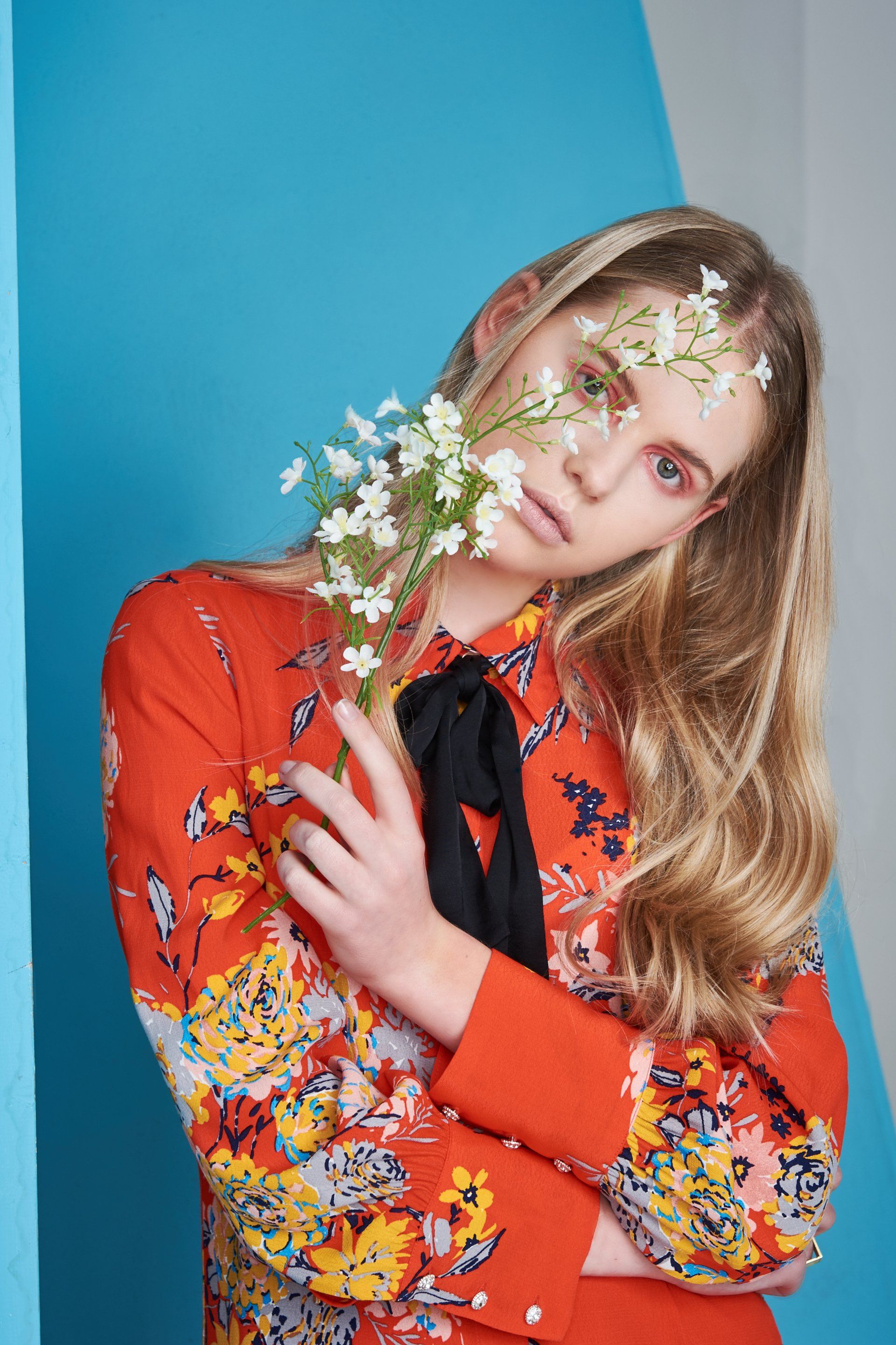 Frau mit blondem Haar in einem roten Hemd mit Blumenmuster, die weiße Blumen vor ihrem Auge hält. 