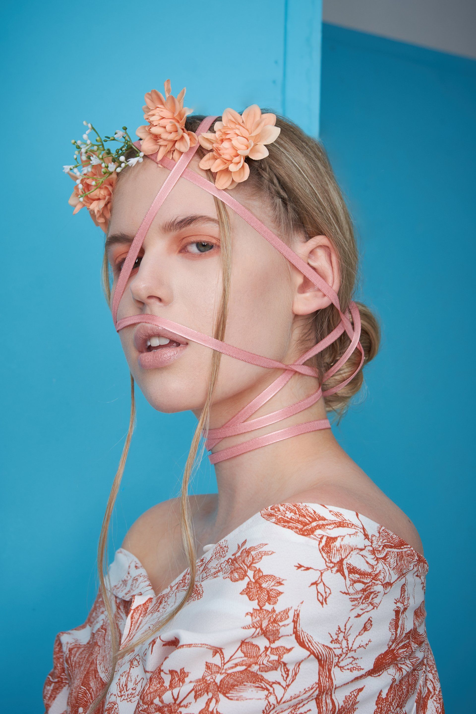 Frau mit Blumenkrone und Band über Gesicht und Hals vor blauem Hintergrund.