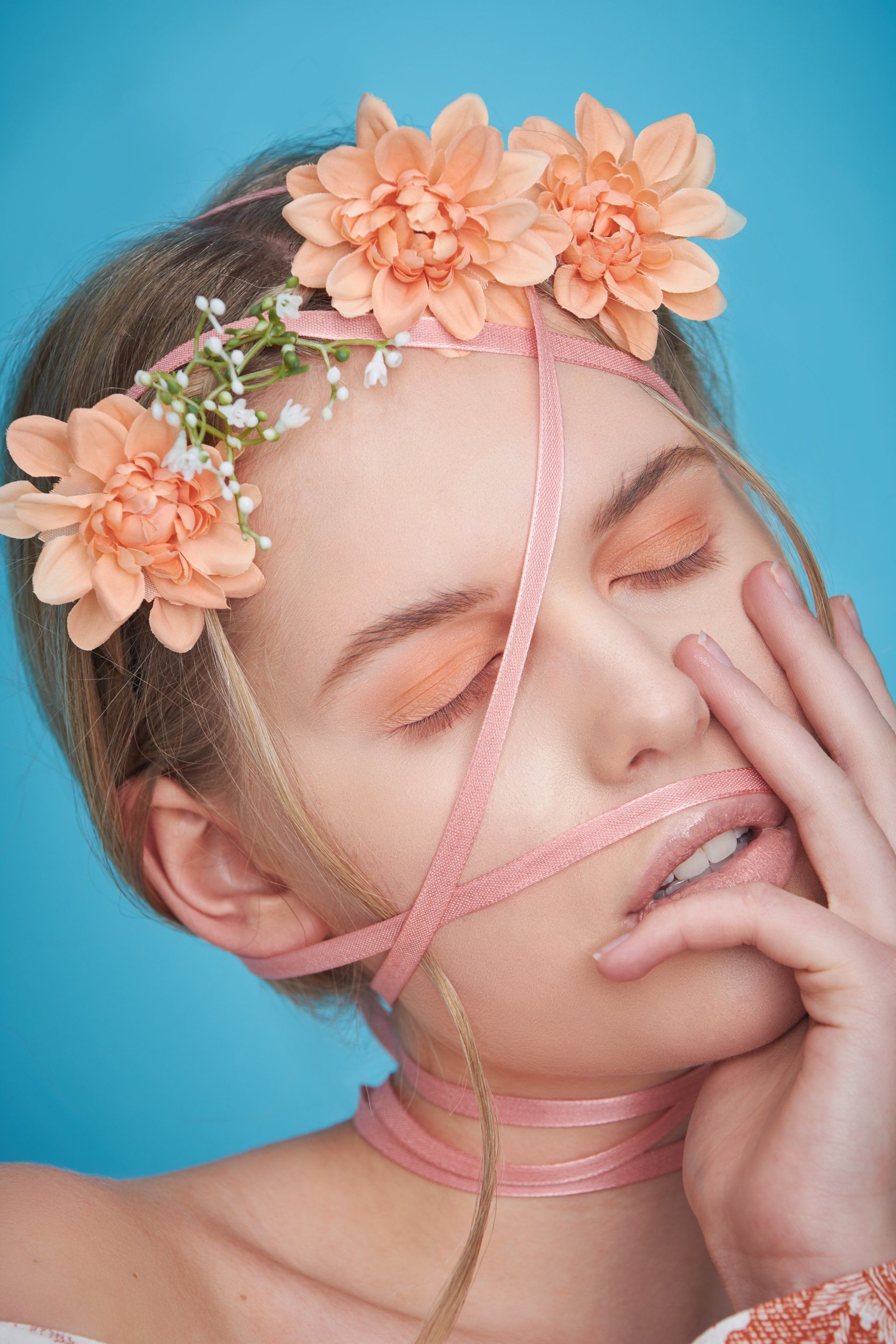 Frau mit pfirsichfarbenem Make-up und Blumenkrone, umwickelt mit rosa Schleife, vor blauem Hintergrund.