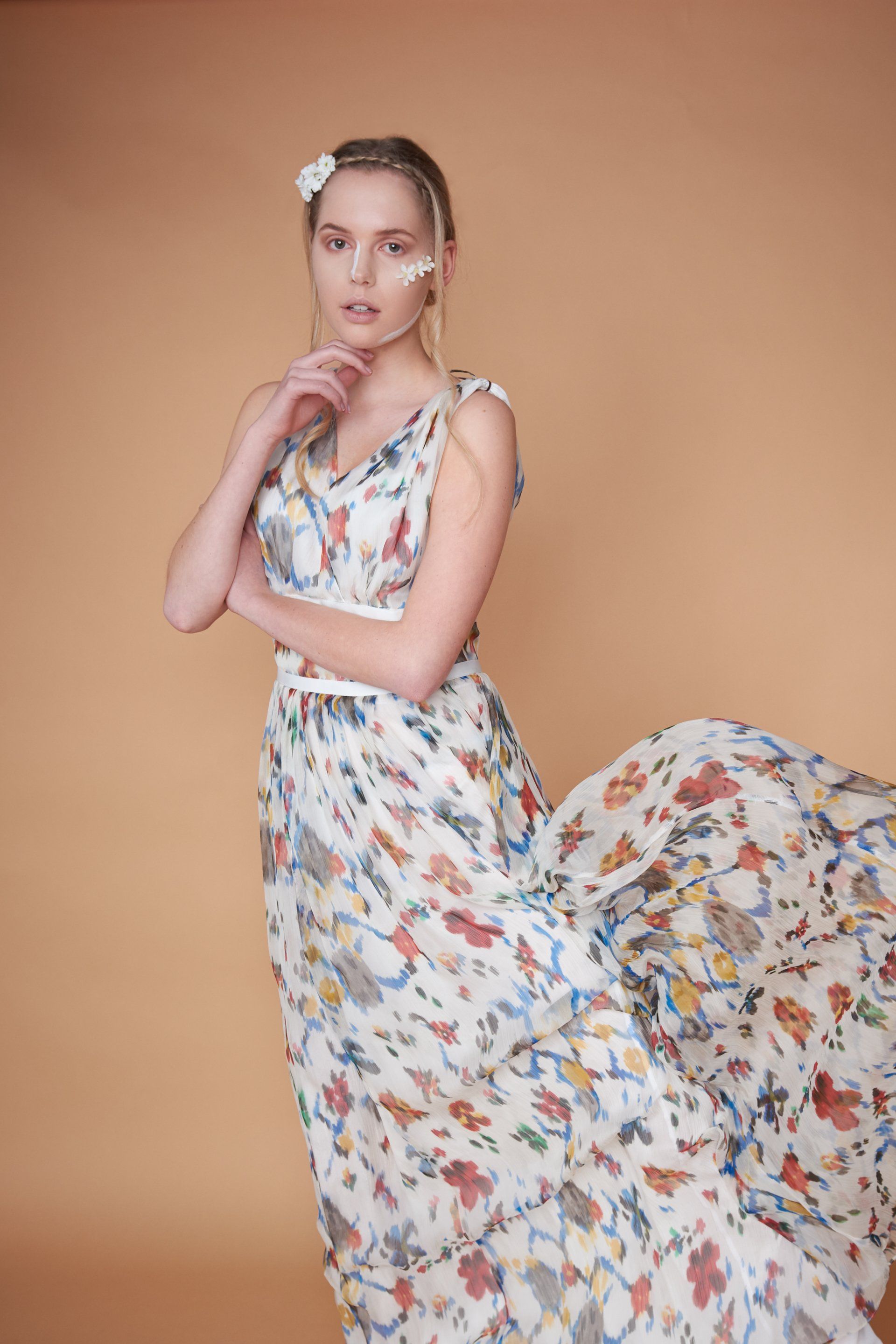 Frau im geblümten Kleid, posiert vor einem pfirsichfarbenen Hintergrund, Hand am Kinn, Blumen im Haar und im Gesicht.