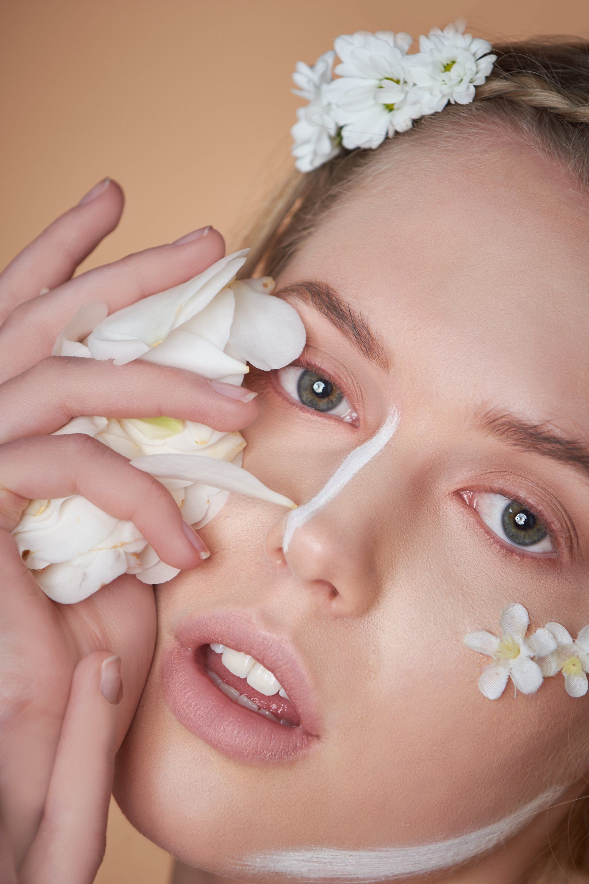 Blonde Frau mit weißen Blumen, Make-up-Linien und offenem Mund. Beiger Hintergrund.