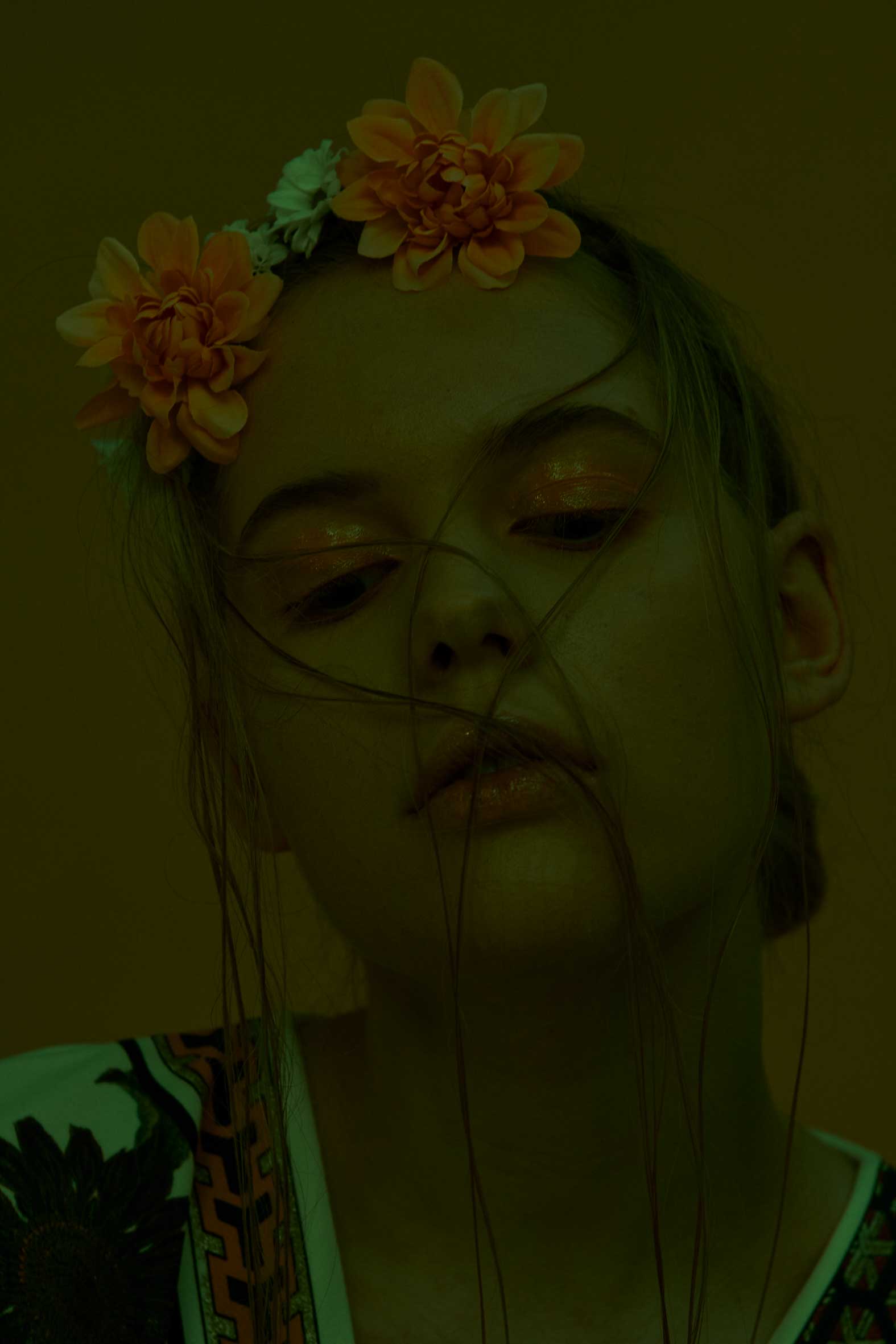 Frau mit Blumenkrone, geschlossenen Augen, Haarsträhnen, goldenem Make-up, gelbem Hintergrund.