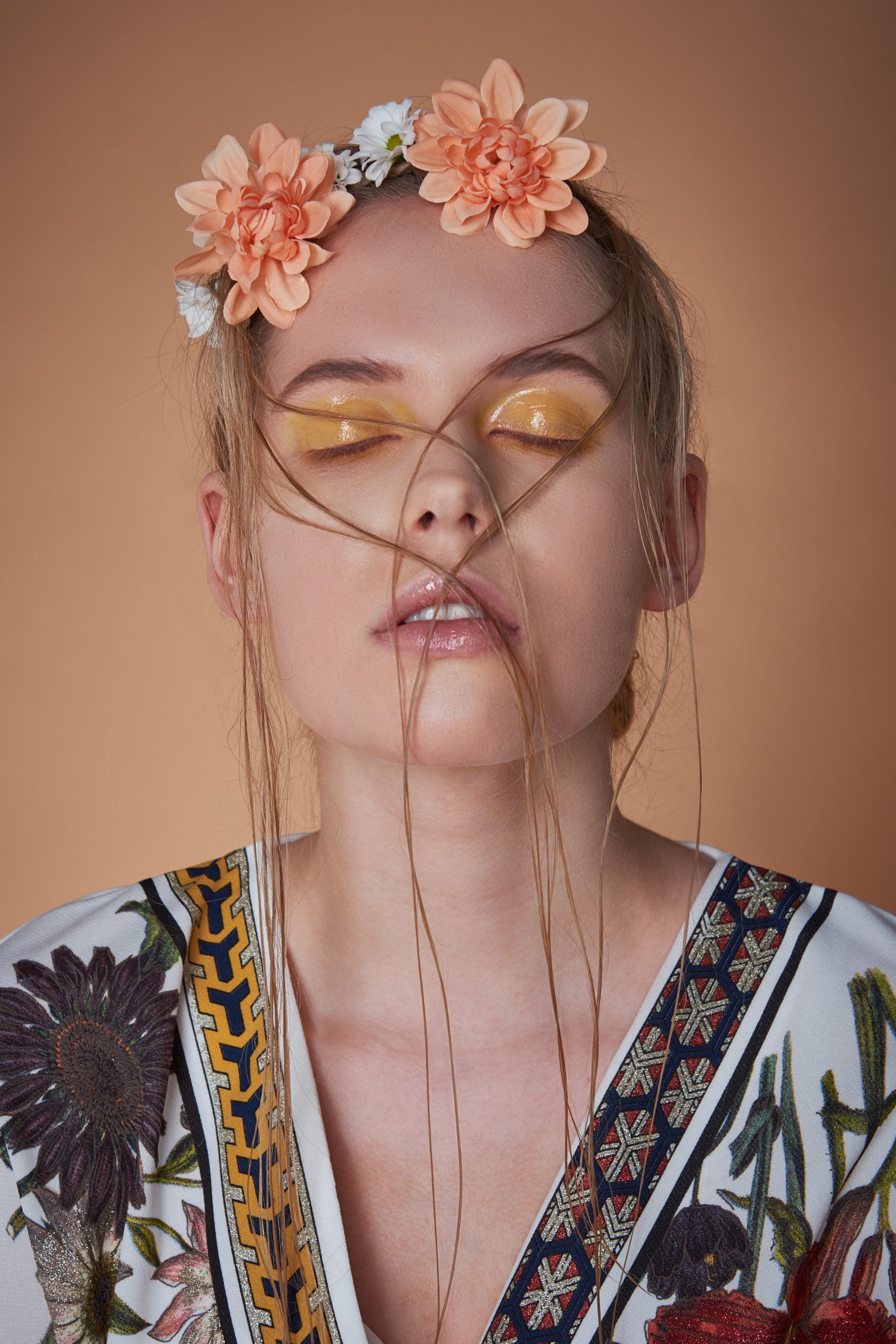 Frau mit Blumenkrone, geschlossenen Augen, goldenem Augen-Make-up und Haarsträhnen im Gesicht vor einem Hintergrund.