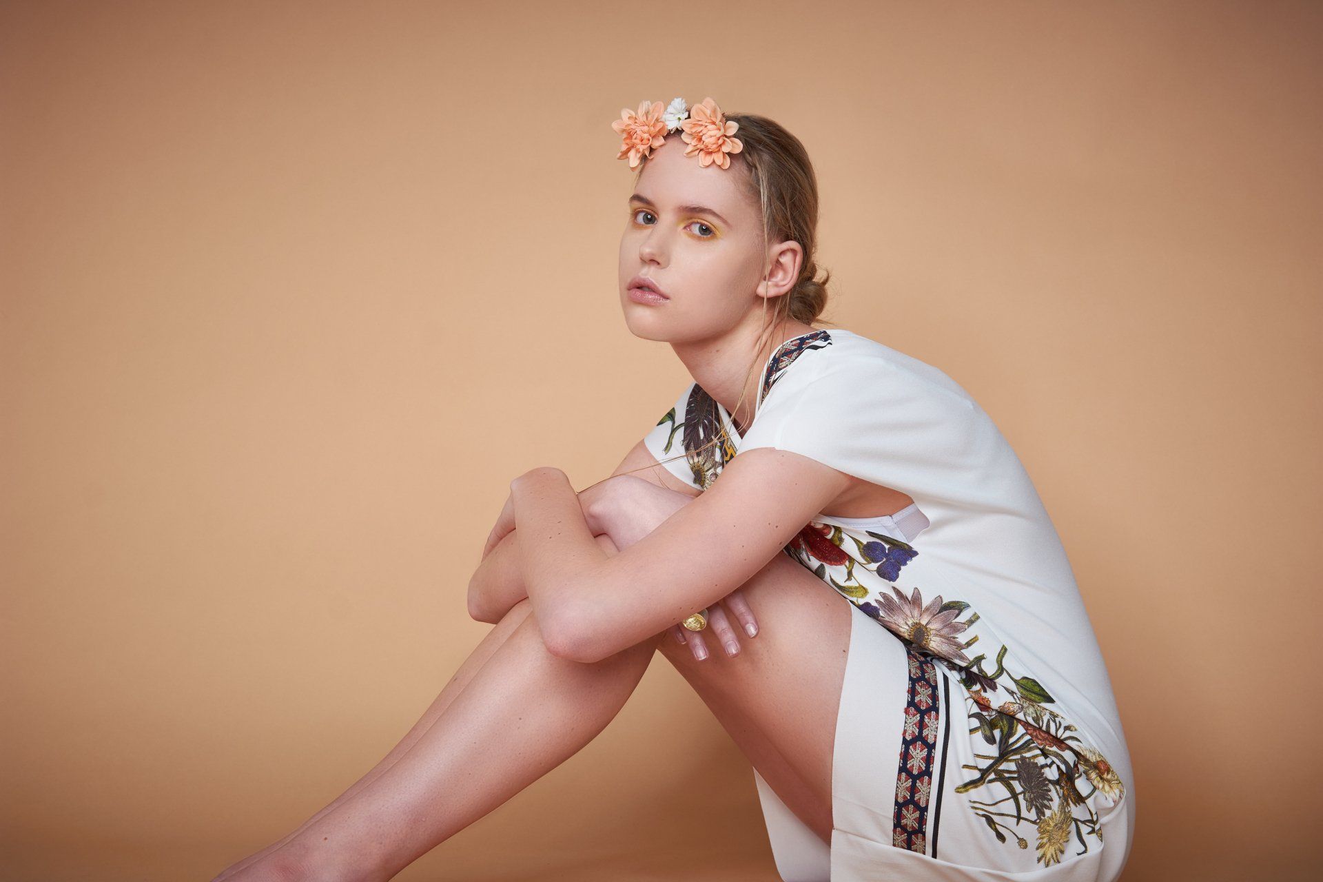 Frau in weißem Kleid und Blumenkrone, lehnt vor einem pfirsichfarbenen Hintergrund und blickt in die Kamera.