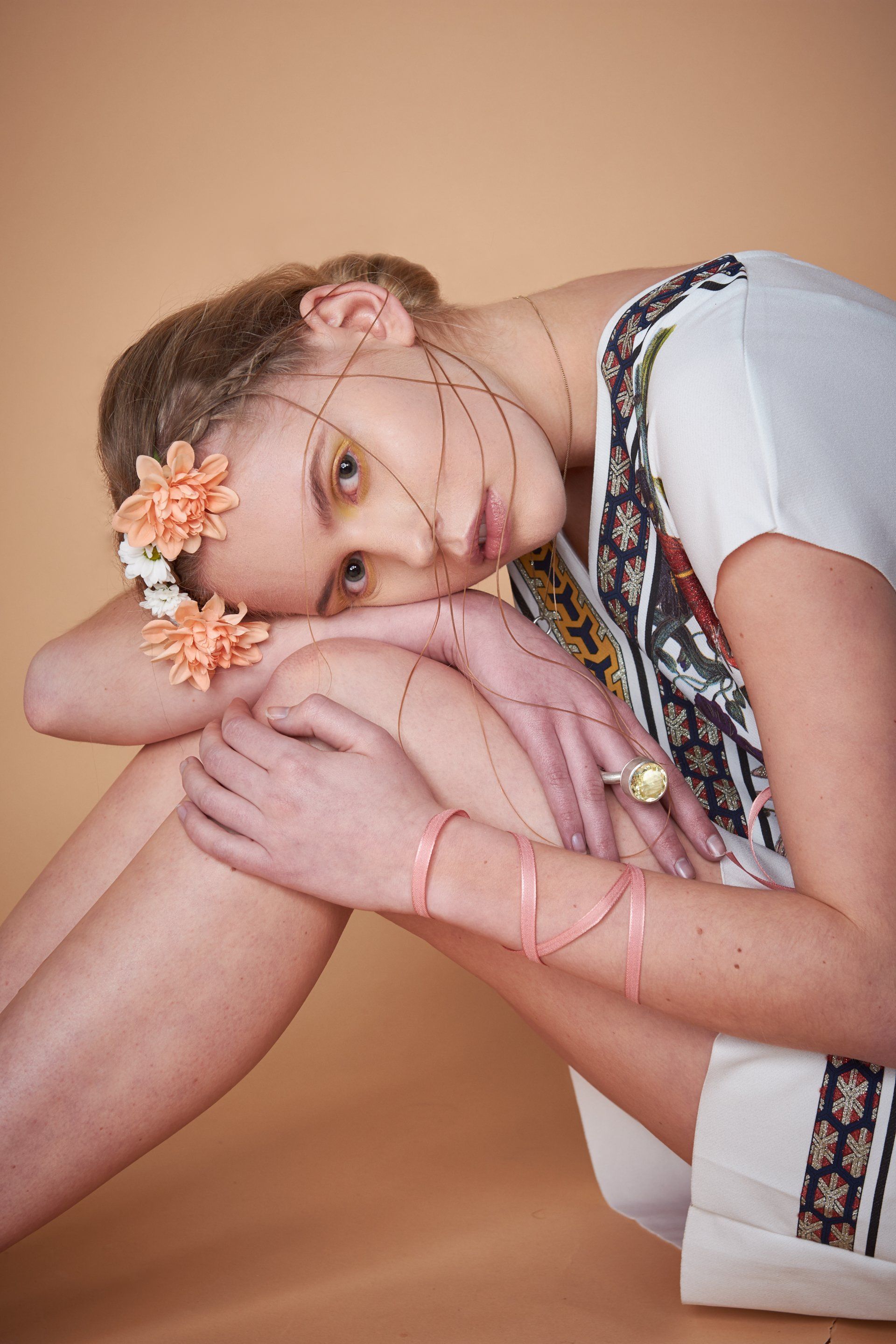 Frau in weißem Oberteil und Blumenkrone, posiert mit String-Art-Linien, Knie an der Brust, vor pfirsichfarbenem Hintergrund.