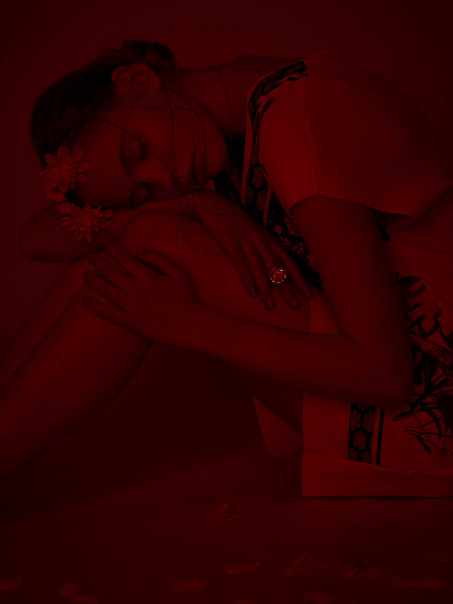 Person zusammengerollt, in rotes Licht getaucht. Augen geschlossen, Arme um die Knie geschlungen. Dunkle Umgebung.