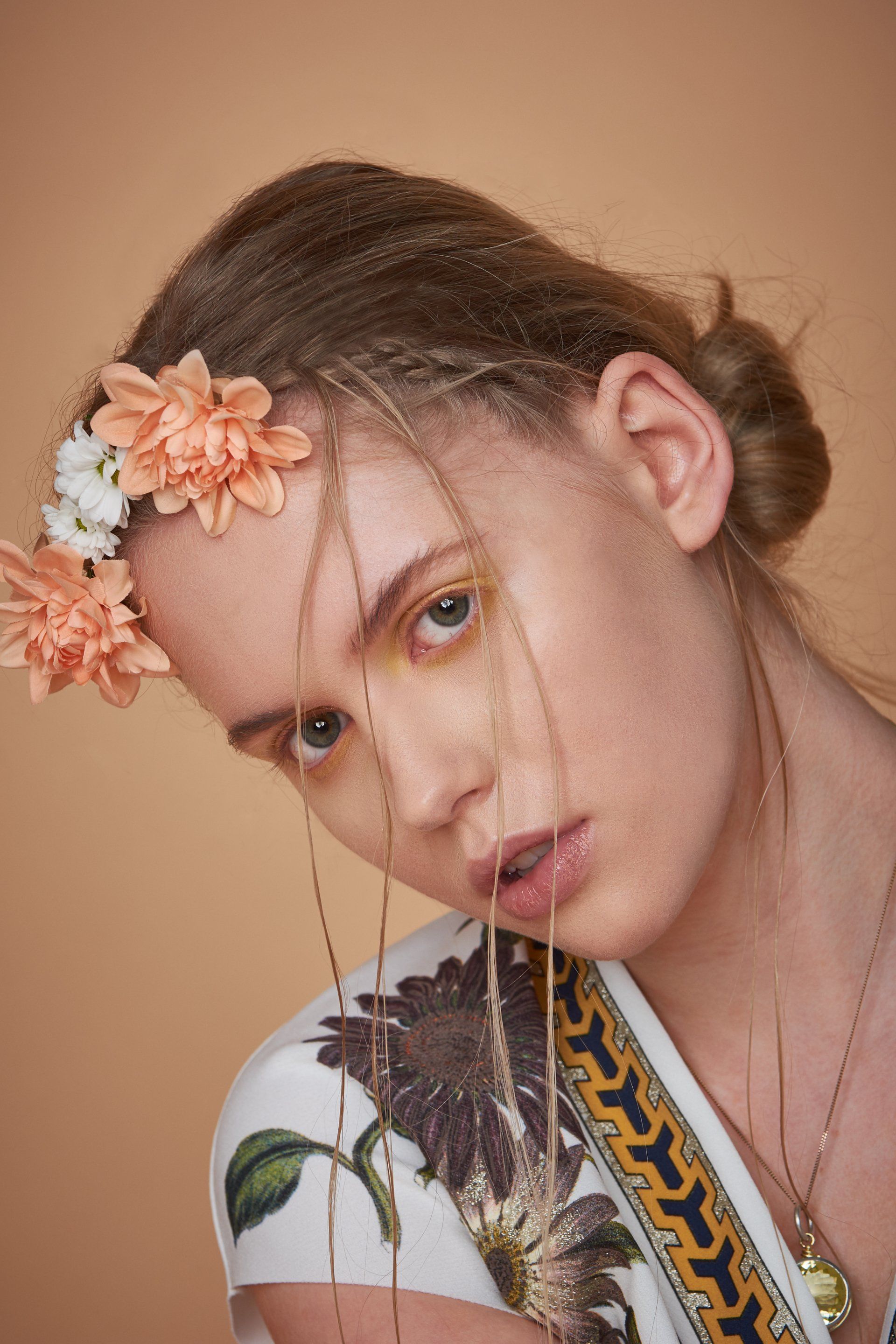 Frau mit Blumenkrone, heller Haut, langen Haaren, geblümtem Kleid und goldenen Akzenten, pfirsichfarbener Hintergrund.