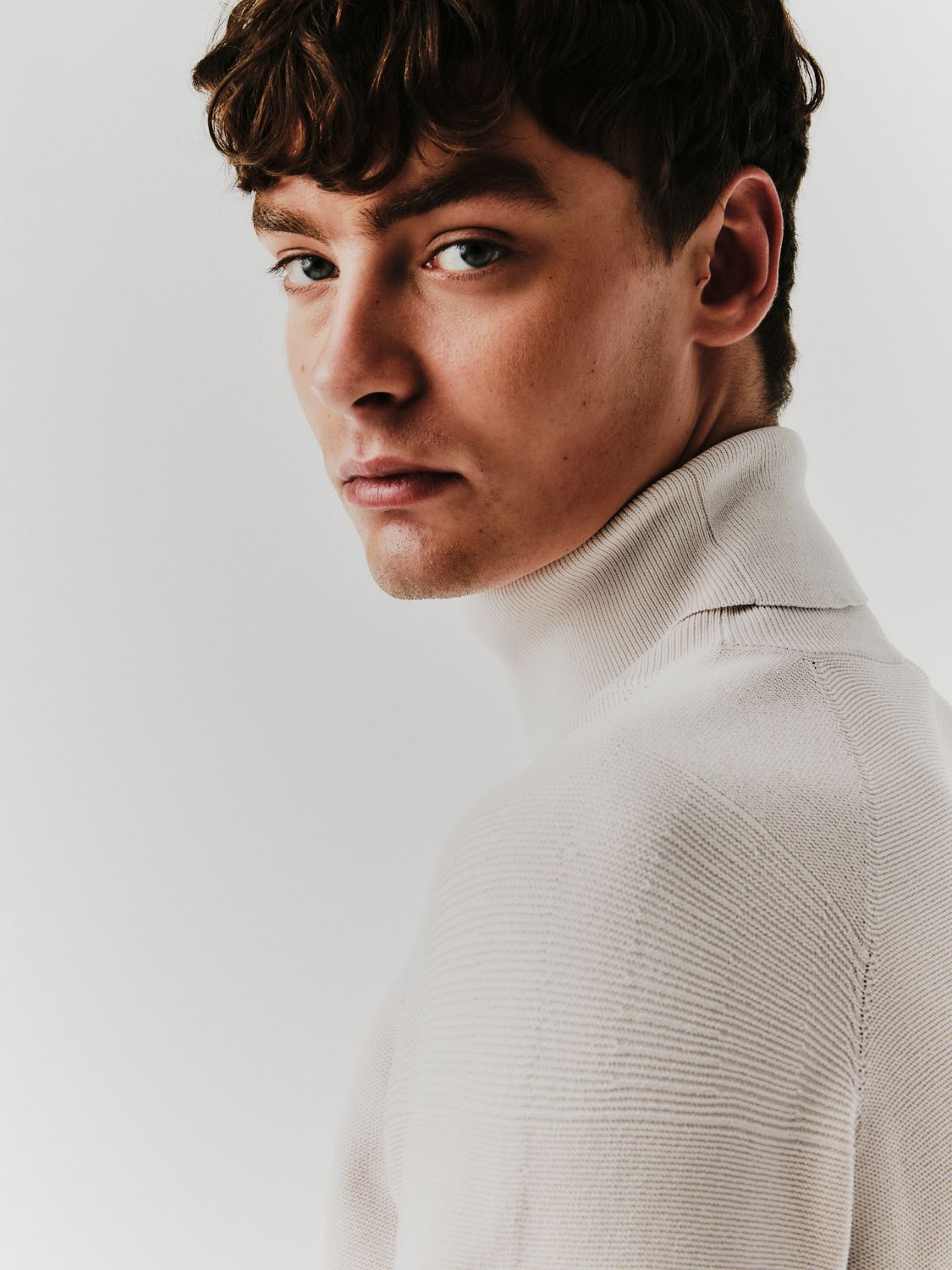 Junger Mann im weißen Rollkragenpullover, der über seine Schulter blickt.