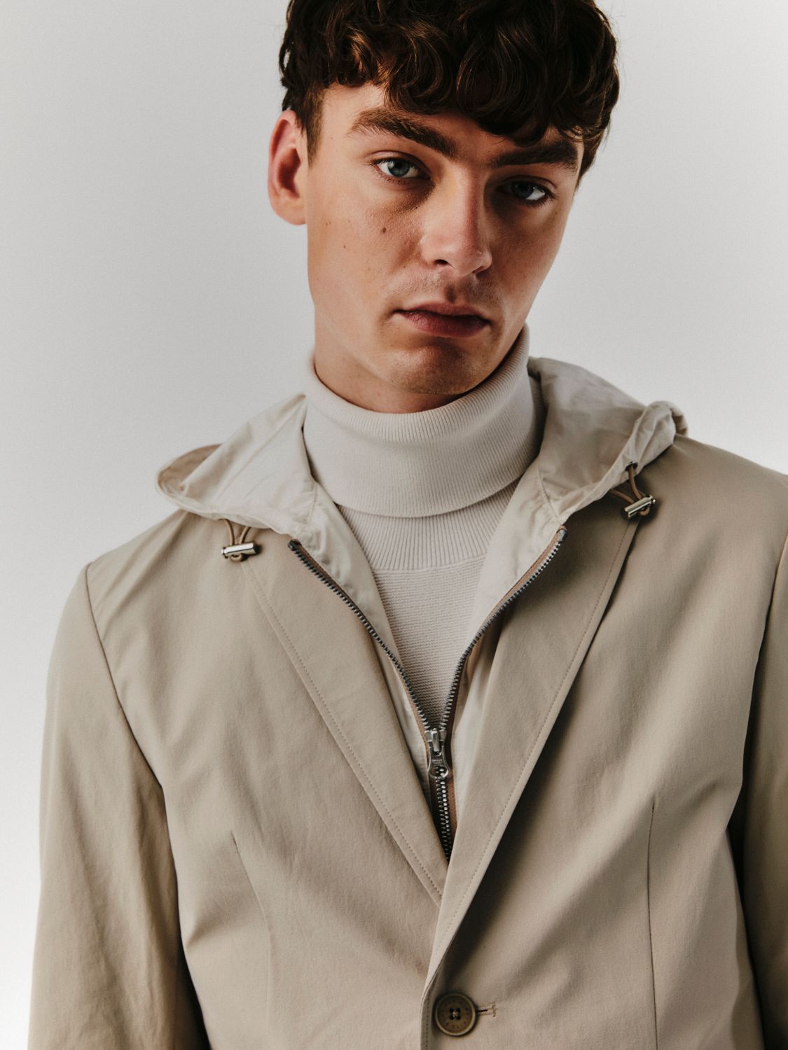 Mann in beiger Jacke mit Kapuze und weißem Rollkragenpullover, blickt in die Kamera.