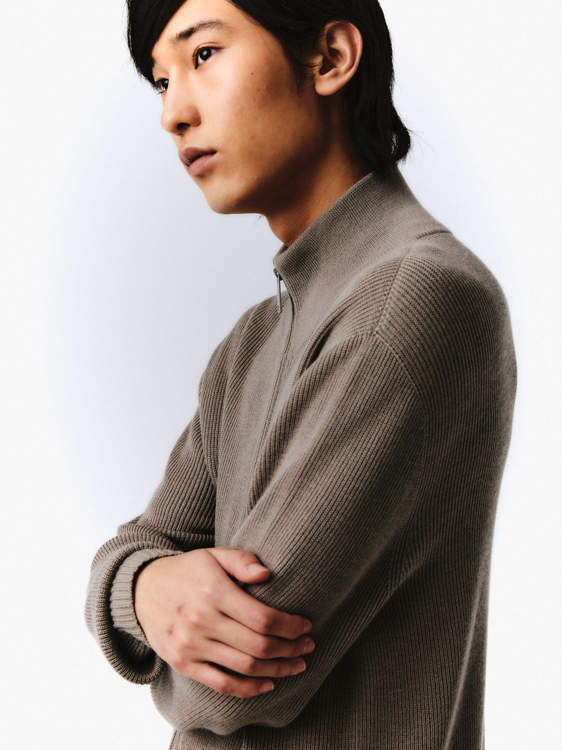 Ein junger asiatischer Mann in einem grauen Pullover, die Arme verschränkt, der Blick nach unten und links gerichtet.